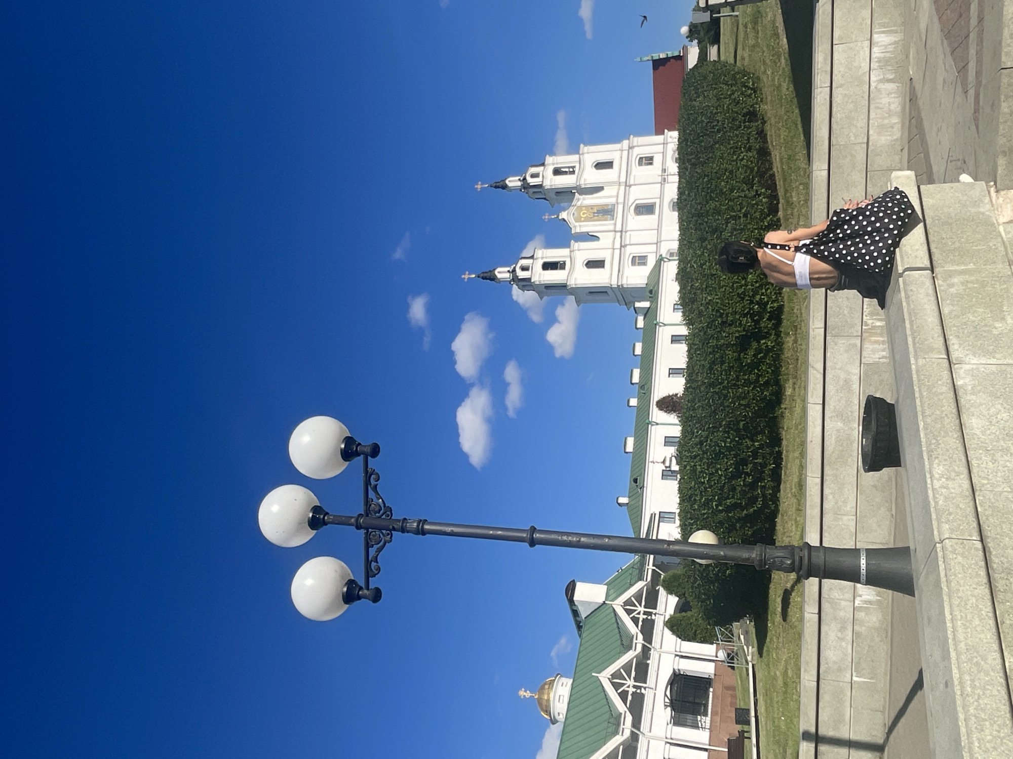 This media file captures a clear, bright, sunny day in an outdoor setting, likely a city square or park, with a large white building featuring multiple spires and ornate architecture in the background. The sky is a vibrant blue with a few scattered white clouds. A lamppost with three white globe-shaped lights stands prominently in the foreground, angled upwards. A person, seen from behind, is sitting on a concrete structure or steps, wearing a black and white polka dot dress and white underwear. They appear to be looking away from the camera, towards the building and the surrounding greenery. The overall impression is one of leisure and a peaceful moment in a historical or religious site, given the architecture of the building. The time of day appears to be midday or early afternoon due to the strong sunlight and shadows. There is no visible text in the image.