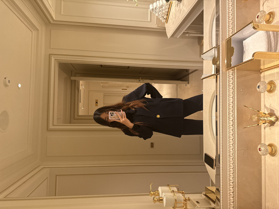 A woman in a dark blazer and matching pants takes a mirror selfie in an opulent bathroom. The bathroom features marble countertops, ornate gold fixtures, a decorative towel rack, and a sink. A long hallway stretches behind the woman, creating an infinite reflection effect. The lighting is warm and inviting. The woman is holding a phone with a floral patterned case.