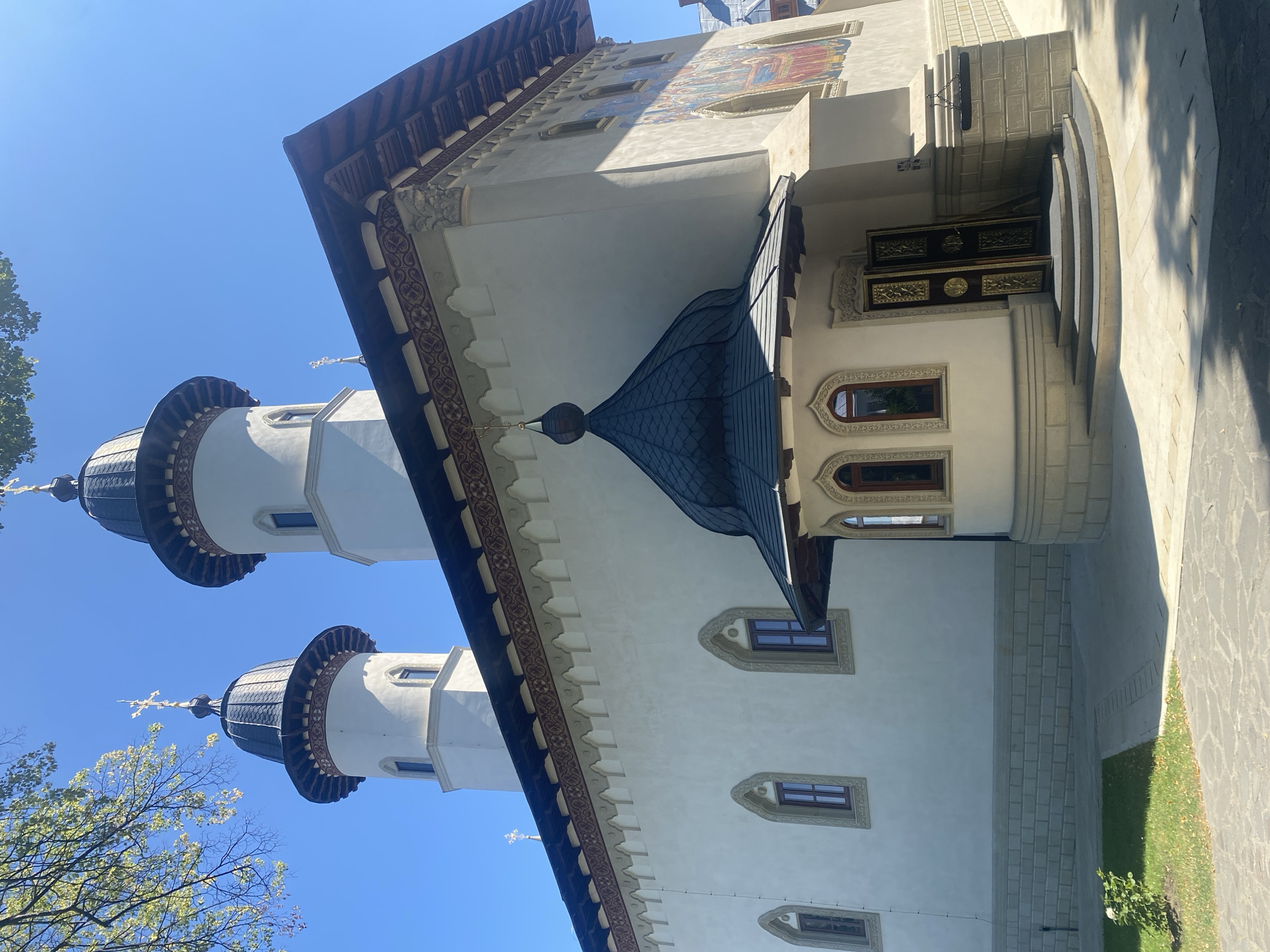 The image shows the exterior of a white church with two prominent dark, domed towers topped with crosses. The church has a tiled roof with ornate trim. There is a smaller, dark, domed entrance structure with arched windows and a decorative golden door. The facade features several arched windows with dark frames and a colorful mural on the right side. The building is situated in an outdoor area with a stone pathway, a grassy area with a small bush, and a clear blue sky. The scene is set in the city of Filioara, Romania, though no people are visible in the photograph.