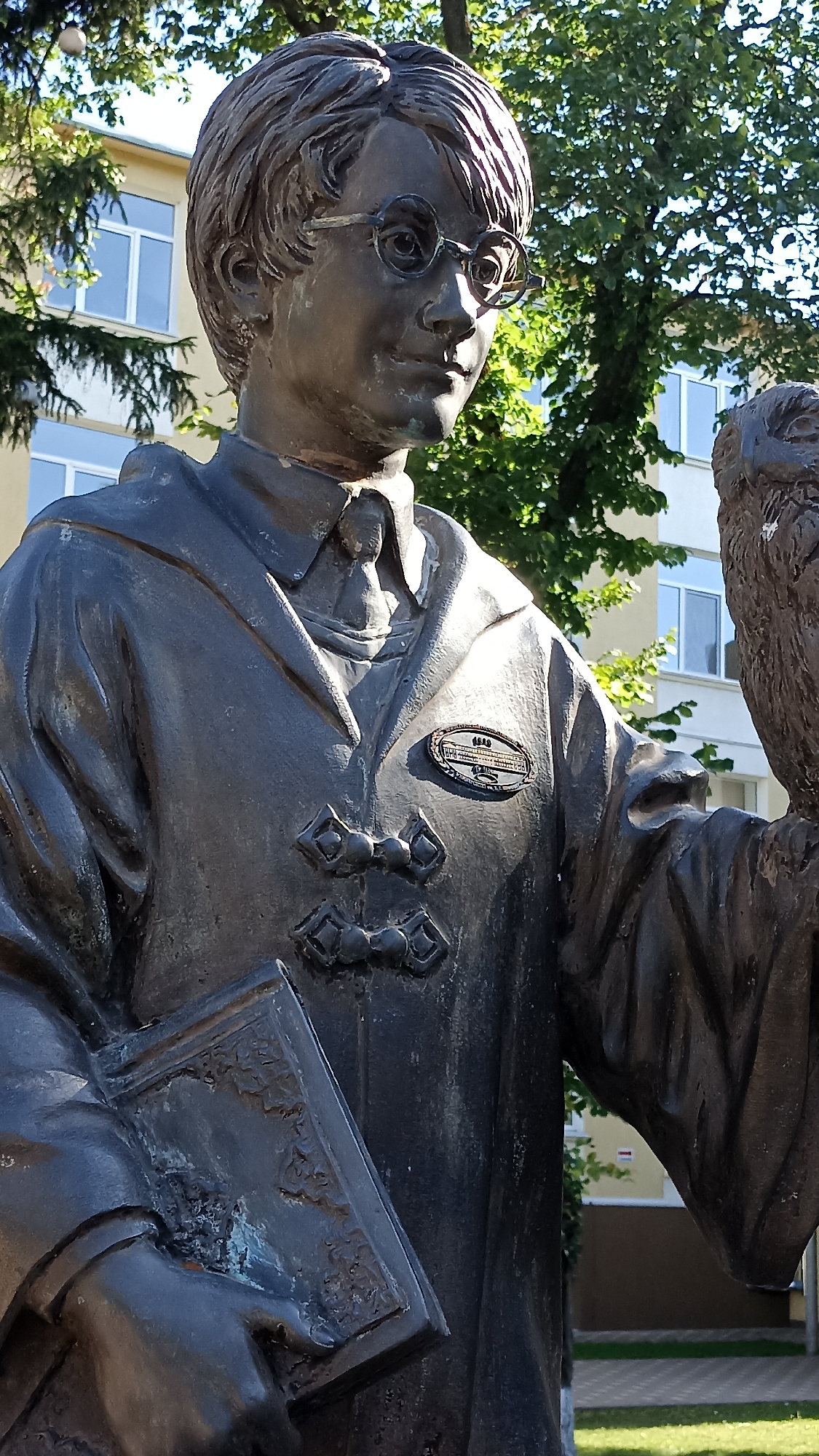 A bronze statue of a young boy wearing glasses and a robe stands outdoors in front of a building. The boy is holding a book in his left hand and an owl in his right. A plaque is affixed to the robe, on the left side of the statue's chest. The statue is located on the grounds of Tiraspol State University in Tiraspol, Moldova. The building in the background appears to be a multi-story educational institution. Trees with green foliage are visible on either side of the statue, and a paved area is in the foreground. The lighting suggests it is daytime.