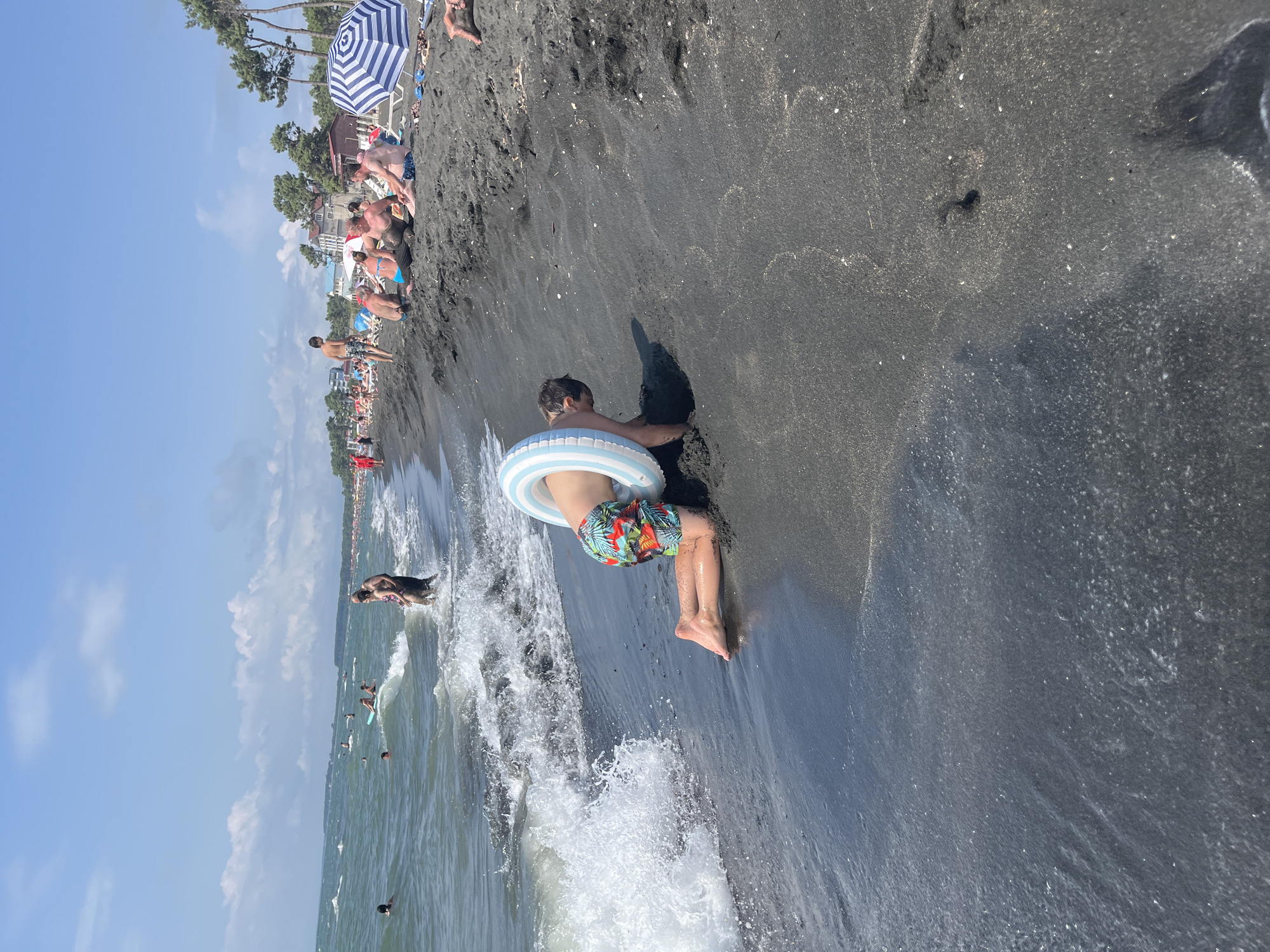 A young boy wearing colorful swim trunks and a striped inflatable ring is kneeling on the wet black sand at the edge of the sea in Ureki, Georgia. His hands are in the sand as he appears to be digging or playing with the wet sand. Waves are breaking near him, creating white foam and spray. In the background, the sea is choppy, and other people are visible in the water and on the beach, some relaxing under umbrellas. The sky is blue with scattered white clouds.