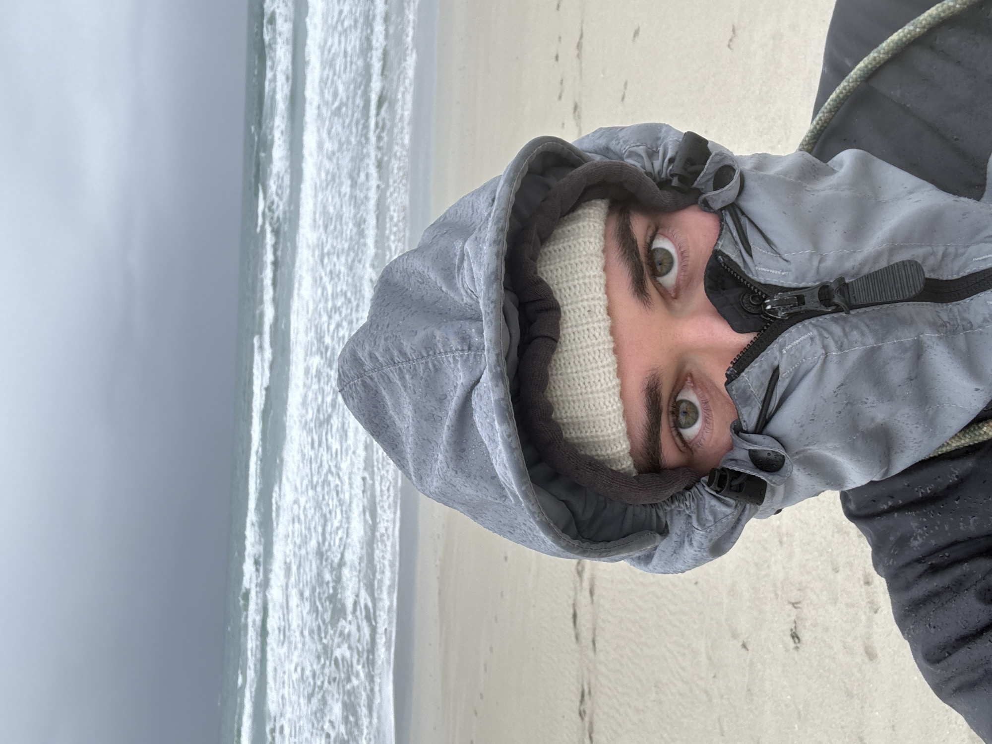 A close-up, rotated image shows a person wearing a light gray hooded jacket and a white knitted beanie, with only their eyes and the top of their face visible. The person is positioned against a backdrop of a beach, with a turbulent ocean and overcast sky in the distance. The scene appears to be taken from a first-person perspective. Footprints are visible on the sandy beach. The location is Unknown, Unknown.