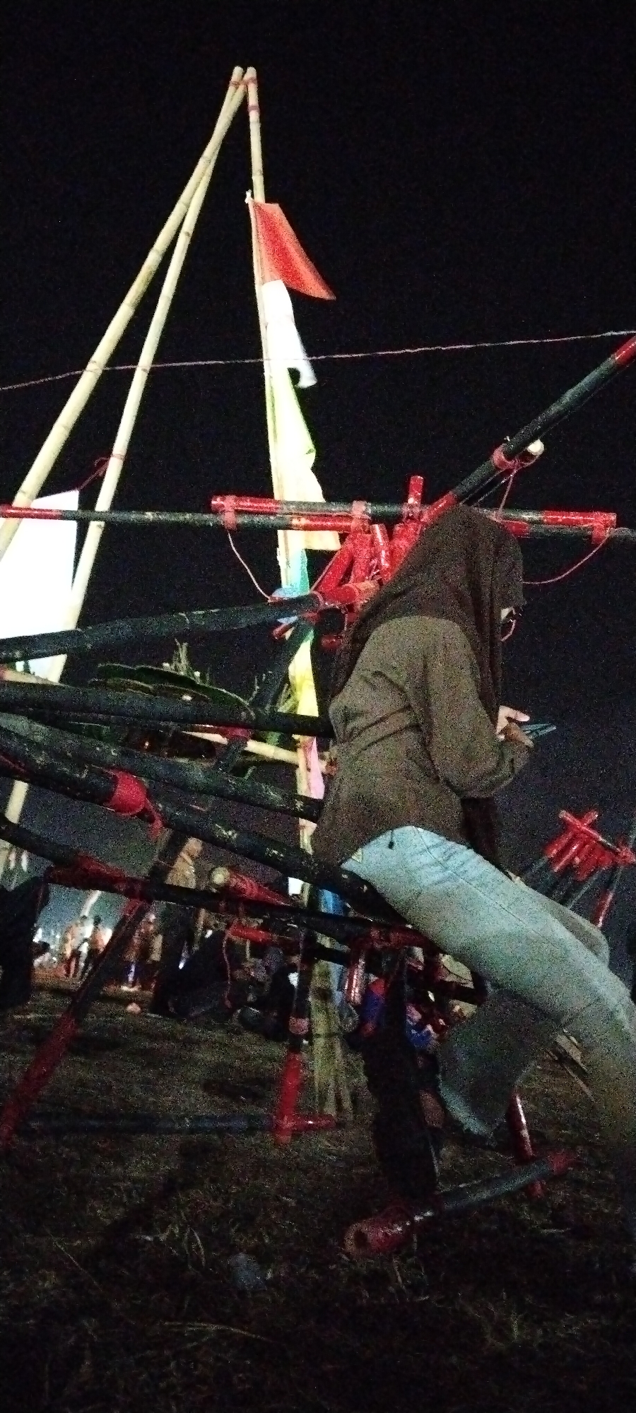 The image depicts an outdoor nighttime scene, possibly at a festival or event, with a person in the foreground. The person is wearing a dark headscarf and a brown jacket over light blue jeans, and appears to be looking at a mobile phone in their hands. They are seated on a structure made of dark, possibly metal, pipes with red accents, which is part of a larger, complex construction. In the background, there are tall bamboo poles with flags attached, including a red and white flag that resembles the Indonesian flag. The ground appears to be dirt or grass, with some scattered debris. The lighting suggests artificial illumination from the event. The overall impression is of a candid snapshot taken during a lively outdoor gathering.
