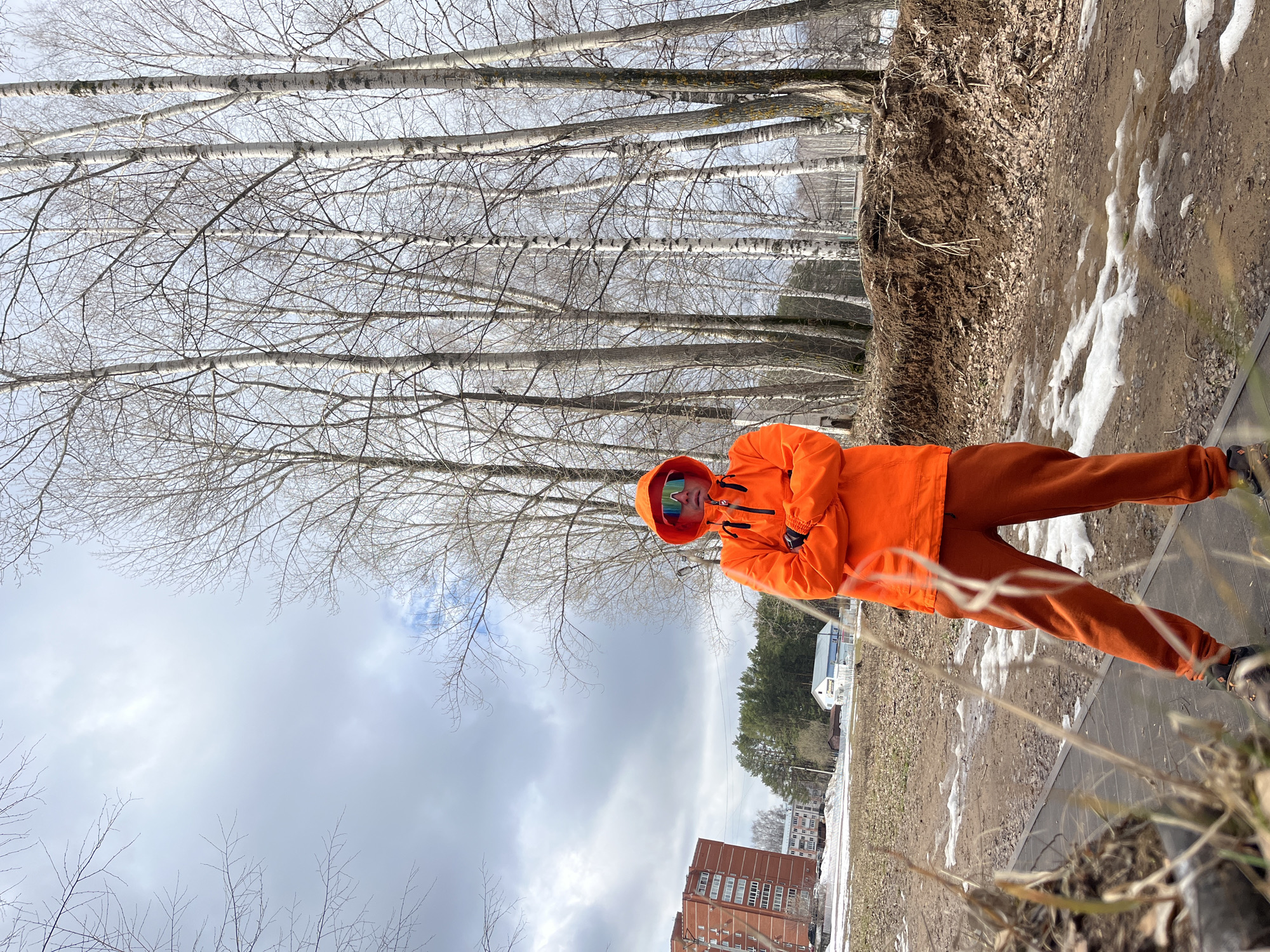 A person in an orange hooded jacket and matching pants stands outdoors near a sidewalk. The location appears to be a park or green space in the city of Chaikovsky, Russia, suggested by the urban landscape elements in the background. The individual is facing the camera with their arms crossed. Behind them are bare trees, and in the distance, there are apartment buildings and a forest. Patches of snow are visible on the ground, indicating it is likely early spring. The sky is overcast.