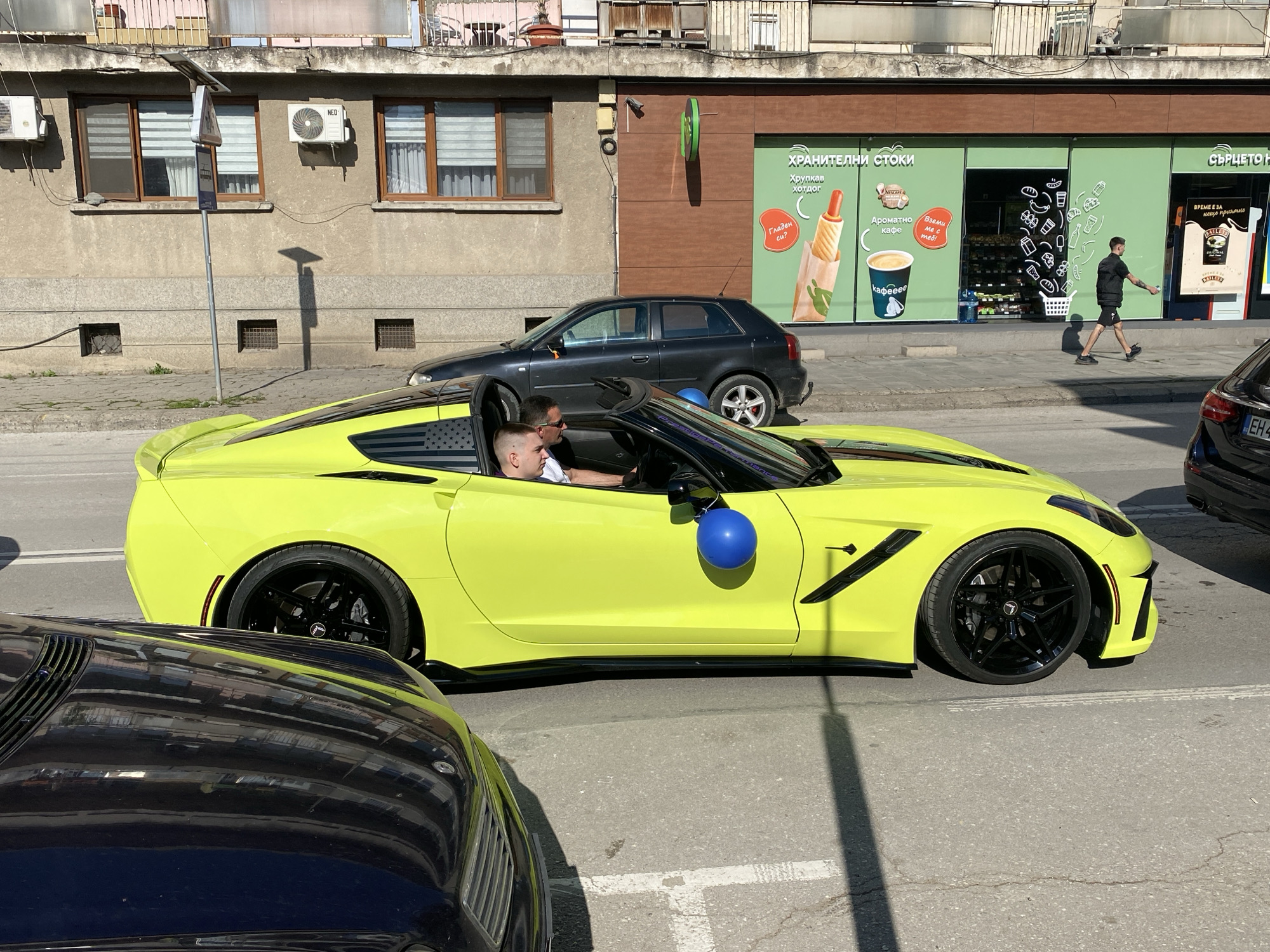 The image captures a scene on a street in the city of Pleven, Bulgaria. A bright yellow Corvette convertible, with American flag decals behind the driver's seat, is parked on the street. Inside the car are two men. One man, visible in the driver's seat, has a shaved head and is wearing a white shirt. The other man has sunglasses on. A blue balloon is tied to the car's side mirror. In the background, there are parked cars, including a dark grey hatchback and a black sedan with license plate "EH."
The building in the background has a green-painted storefront with windows displaying advertisements for food and beverages. A man in a black outfit is walking on the sidewalk in front of the store. The street is paved and marked with white lines. The time of day seems to be daylight with sunny conditions.