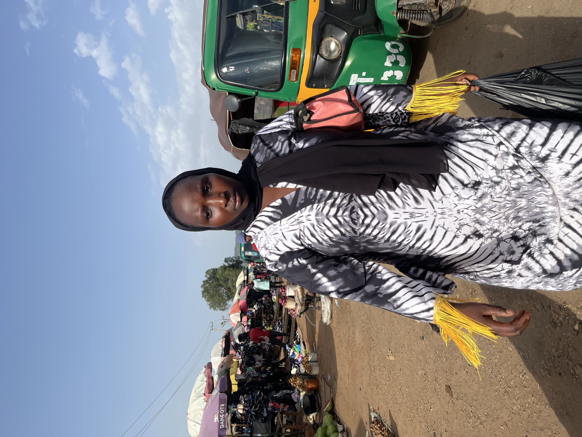 A woman stands in the foreground, facing the camera. She wears a black headscarf and a black and white patterned dress with yellow fringe on the sleeves. She is holding a pink bag over her left shoulder and a black plastic bag in her right hand. Behind her, a green and yellow auto-rickshaw is parked. The background shows an outdoor market scene with various stalls, umbrellas, and people. The ground is dirt and sand. The sky is blue with scattered clouds. The image appears to be taken in the city of Rafin Par, Nigeria.
