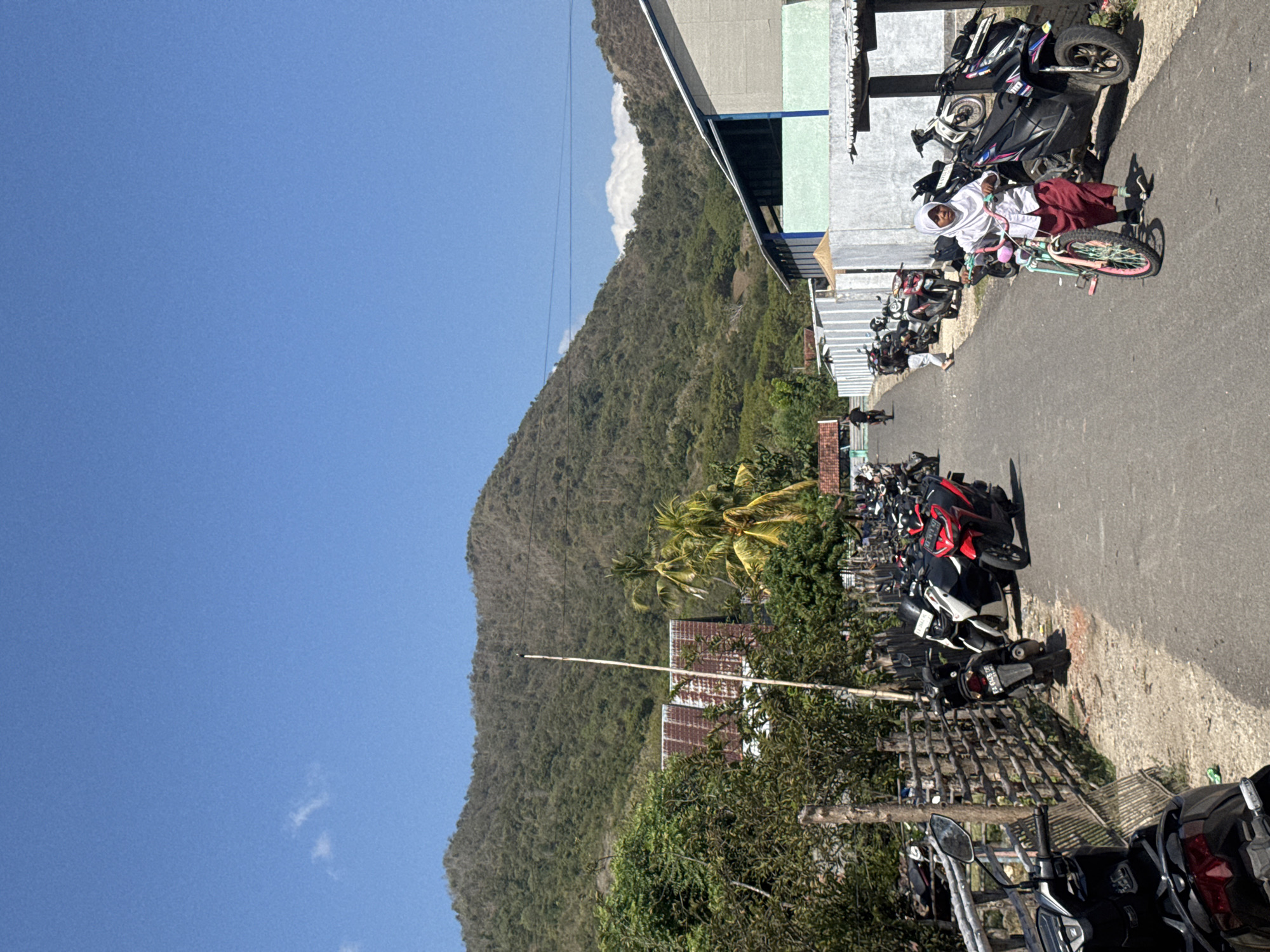In Kertasari, Indonesia, a street scene unfolds under a clear blue sky with scattered clouds. A paved road runs through the frame, lined with parked motorcycles on both sides. On the left, a rustic wooden fence separates the road from a patch of greenery and trees. Behind the fence, a building with a rusted metal roof is visible, along with palm trees. Further back, a large, green, tree-covered mountain dominates the landscape.
On the right side of the road, a modern building with a light-colored facade and a blue trim runs along the edge. Several motorcycles are parked in front of this building. In the foreground on the right, a young person wearing a white hijab and a maroon skirt is walking alongside a pink bicycle. Their gaze is directed forward, and they appear to be pushing the bicycle. Further down the road, a few other individuals are visible walking or on motorcycles, and more motorcycles are parked in the distance. The overall impression is a sunny day in a semi-rural or suburban area.