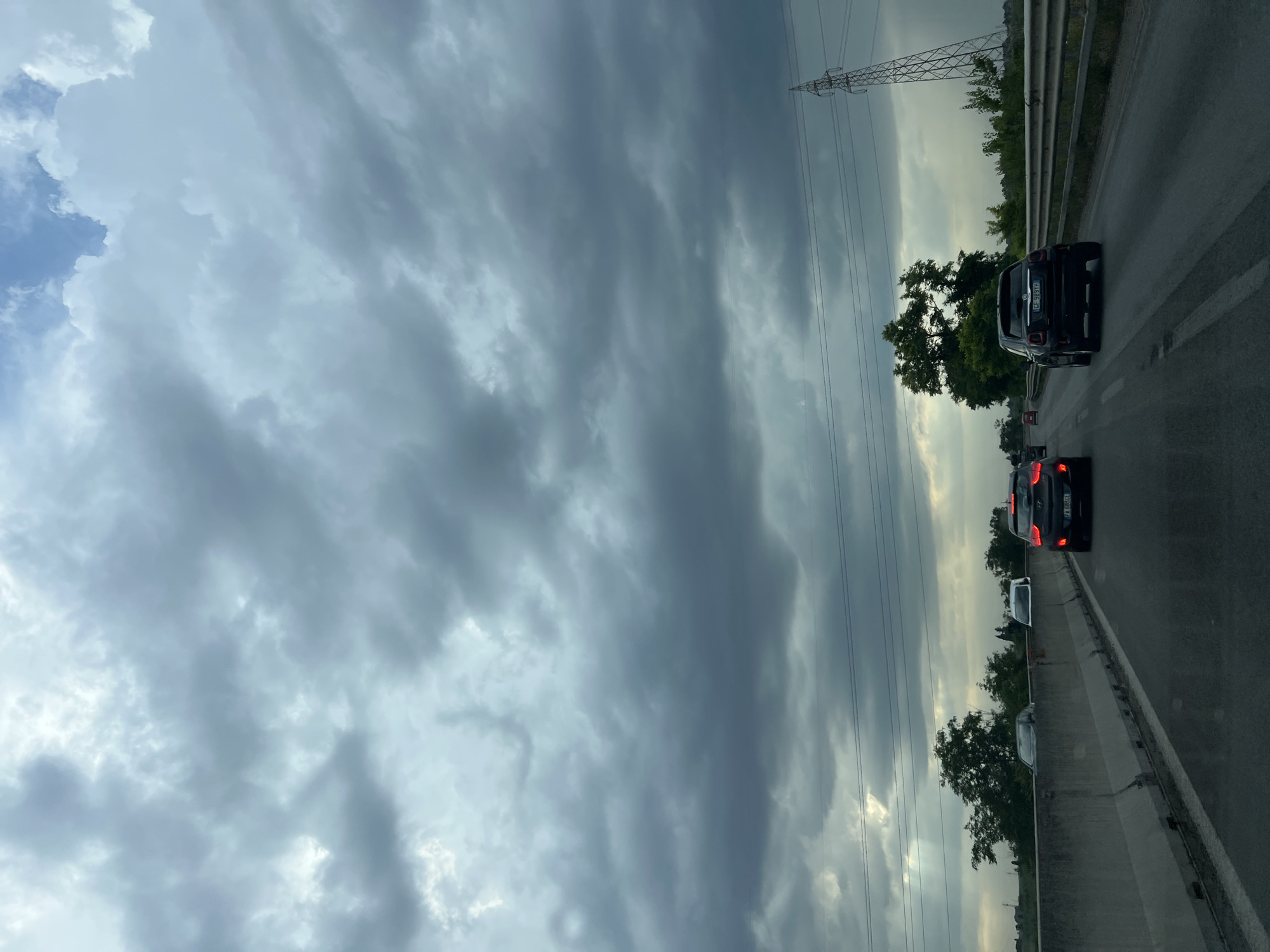 A vertical, eye-level, wide shot captures a view from a vehicle traveling on a highway, looking forward. The sky is filled with dramatic, dark gray clouds, suggesting an impending storm. The road ahead is asphalt with a white dividing line, and on the right, a metal guardrail separates the highway from a grassy embankment. A large, dark gray concrete barrier runs along the left side of the road. Several cars are visible on the highway, including a dark colored Fiat 500 in the foreground on the right lane, and a red car further ahead in the left lane. Beyond the guardrail, there are trees and vegetation. In the background, a tall, lattice-like electrical pylon stands prominently on the right. The overall scene conveys a sense of travel under brooding, overcast skies.