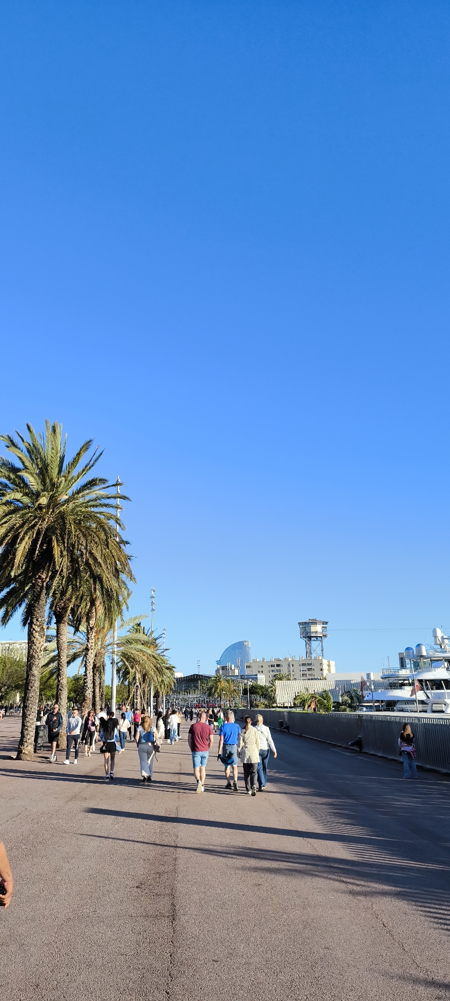 The image depicts a scene along a promenade in Barcelona, Spain. The environment is clear and sunny, with a bright blue sky dominating the upper portion of the frame.
A crowd of people is walking along a wide, paved pathway. The people are diverse in age and attire, walking in various directions. Palm trees line the left side of the path.
In the background, buildings and structures are visible, including a distinctive sail-shaped building (Hotel W Barcelona). Yachts are docked further in the background on the right. A tall observation tower is seen beyond the structures. A metal fence is present on the far right.