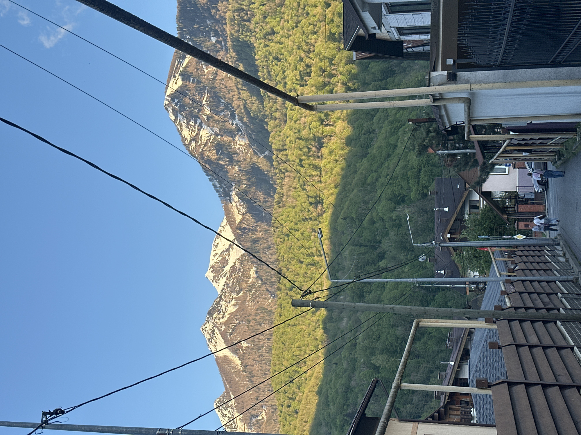 The image depicts a street scene in Krasnaya Polyana, Russia, with a large mountain range dominating the background. The mountains are partially covered in snow, with dense green forest covering the lower slopes. In the foreground are buildings and infrastructure characteristic of a small town, including utility poles and power lines that crisscross the sky. The road is visible, with at least two people walking along the side.