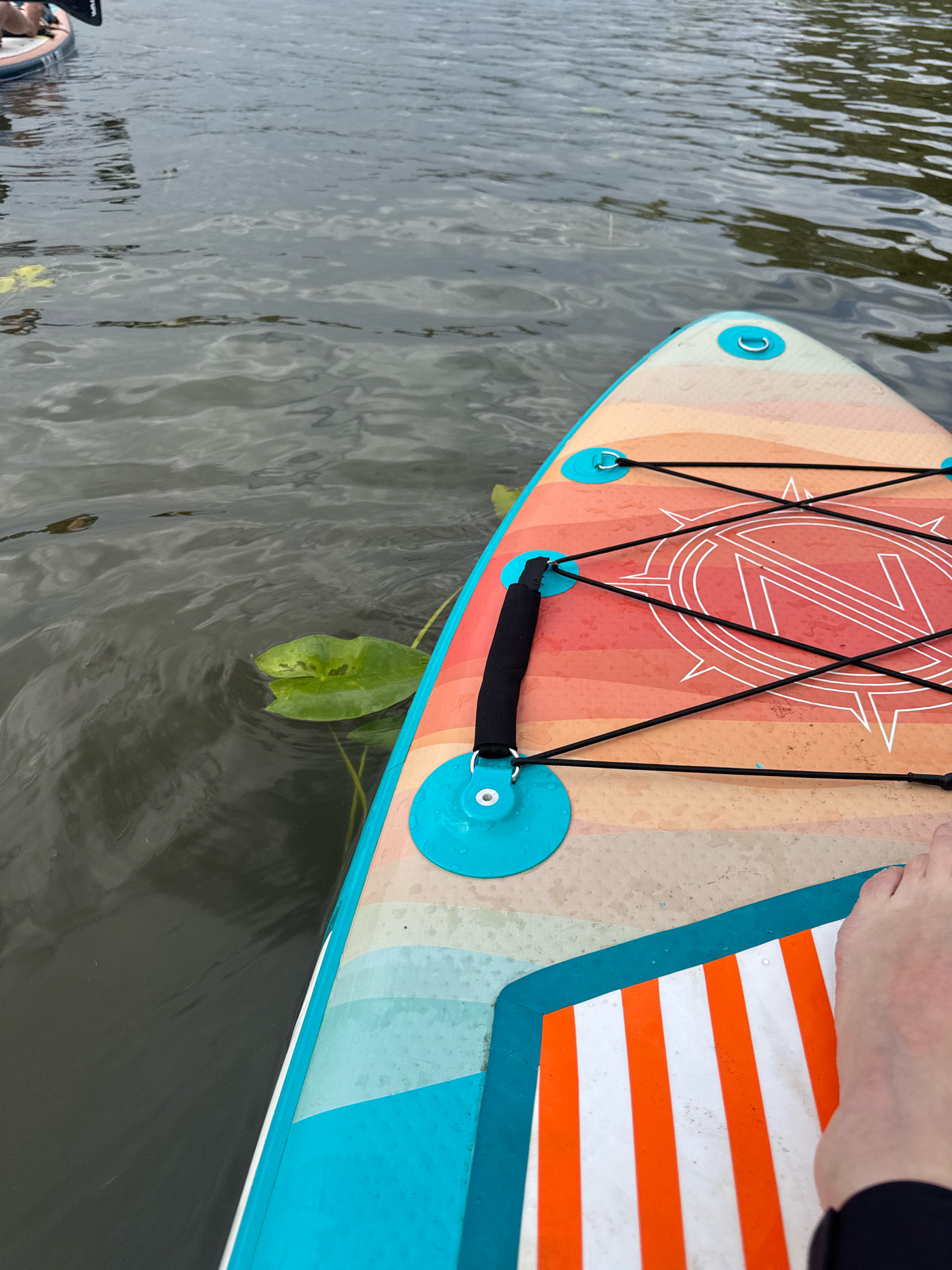 The image shows a first-person perspective of someone on a paddleboard on a body of water. The paddleboard is colorful with orange, yellow, and blue stripes, and features a compass-like design in the center. There is a black bungee cord system for securing items. A single lily pad is visible in the water to the left of the paddleboard. The water appears murky with ripples. The location of Ufa, Russia, is not discernible from the image.