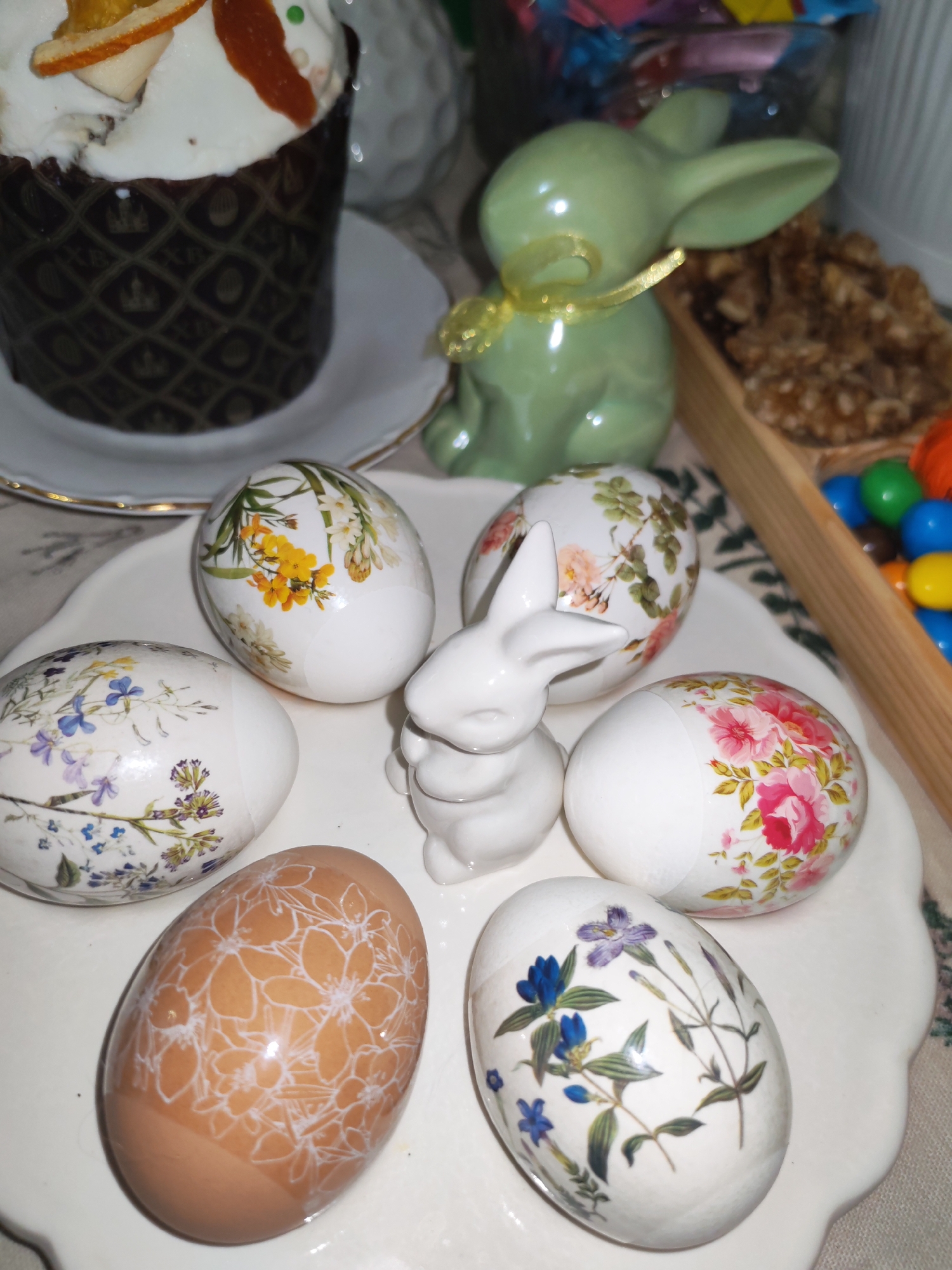 A still life composition featuring Easter decorations. On a white plate with a scalloped edge, there are six decorated Easter eggs and a small white ceramic bunny figurine. The eggs have floral decals in various colors, including yellow, pink, blue, and white. One brown egg features a white floral pattern. A light green ceramic bunny with a yellow ribbon is positioned behind the eggs. To the left, partially visible, is an Easter cake decorated with icing and candied fruit on a white plate. To the right, a wooden tray contains walnuts and colorful candies. The background is slightly blurred, showing other objects like jars and possibly other Easter treats. The setting appears to be indoors, likely in a home. There is no information available in the image to suggest it was taken in Izhevsk, Russia.