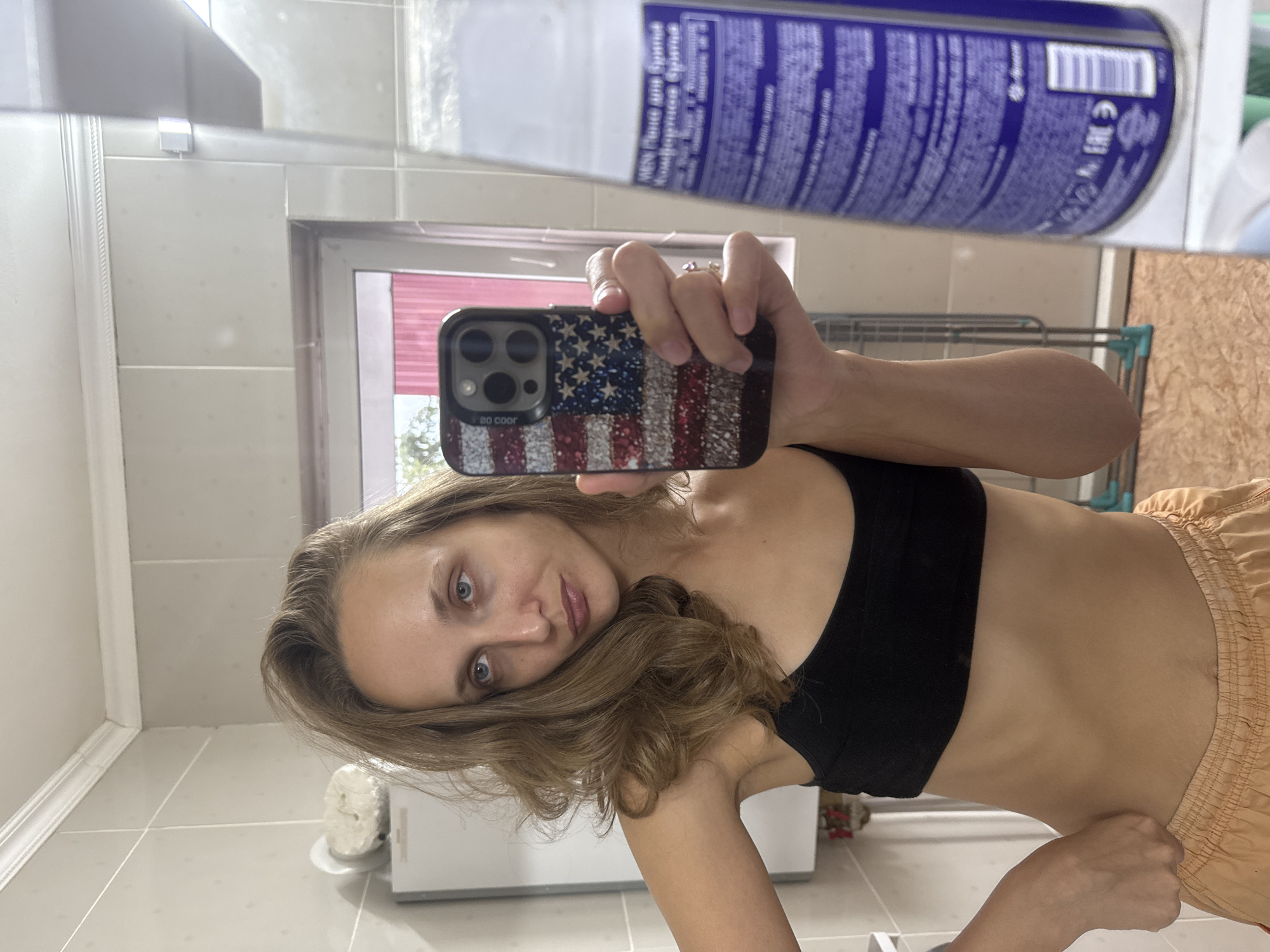 A woman is taking a mirror selfie in a bathroom. She is wearing a black sports bra and orange shorts. She is holding a phone with an American flag case. The bathroom has tiled walls and a mirror. A bottle of shaving cream is visible in the reflection. The background shows a window with pink blinds and a clothes drying rack. The image was taken in Kuyeda, Russia.