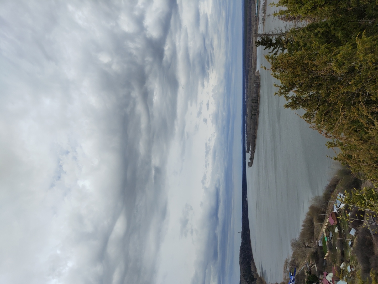 The image depicts a view of the Volga River in Ольховка, Russia from an elevated position. The river stretches into the distance under a cloudy sky. The river is bordered by trees and vegetation, and houses are situated along the bank. The photograph is taken from an angle, and the horizon is not level.