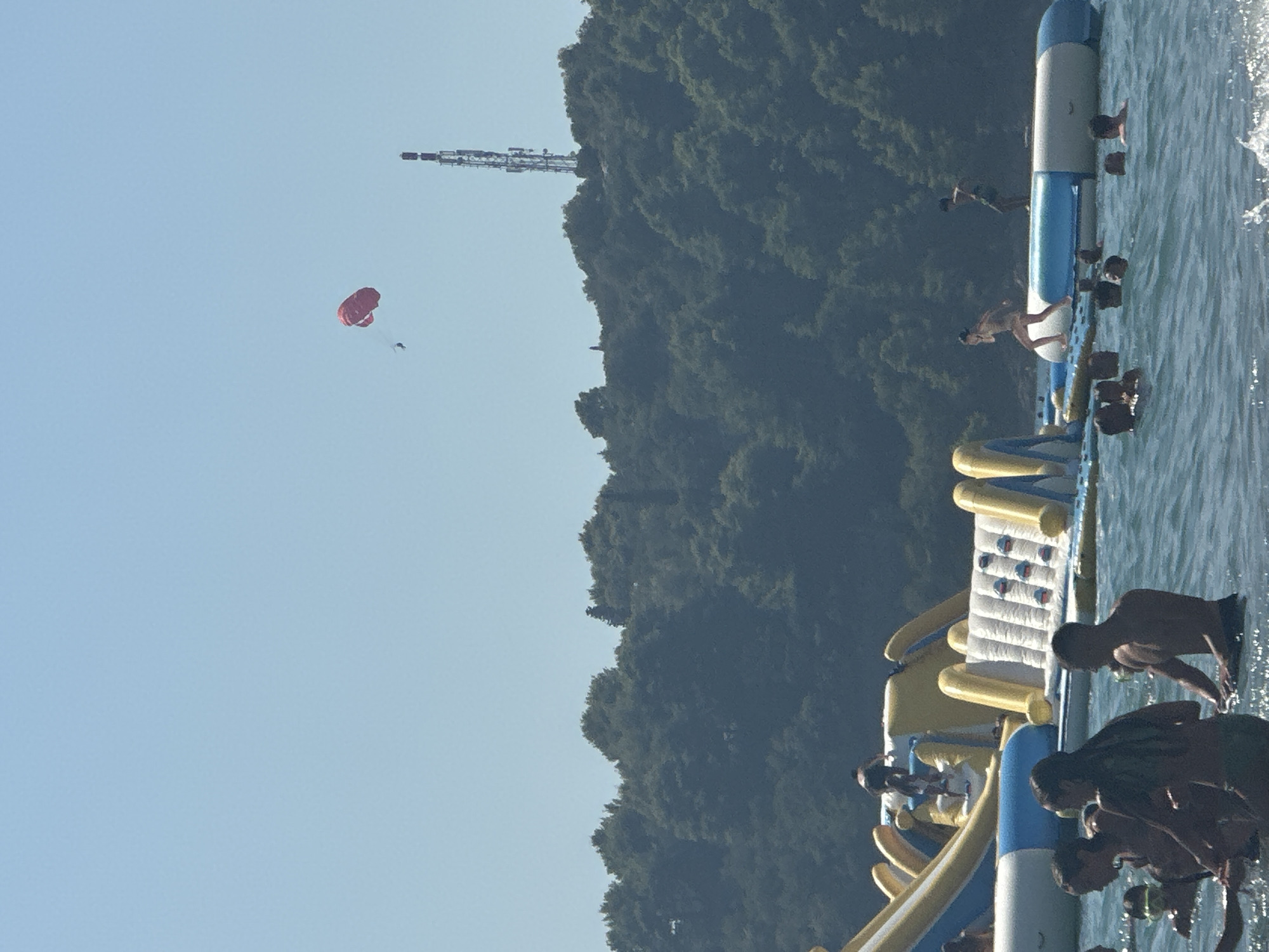 In Boreti, Montenegro, a bright, sunny day is depicted with people enjoying recreational activities on the water. A large inflatable water park, featuring slides and climbing walls, dominates the mid-ground. Several individuals are seen interacting with the water park, with some climbing or preparing to slide, and others swimming in the clear blue water. In the background, a dense, green forest covers a hillside, topped by a tall communication tower. High above, a person is visible paragliding with a red parachute against a clear, pale blue sky. The scene suggests a lively atmosphere at a popular summer resort.