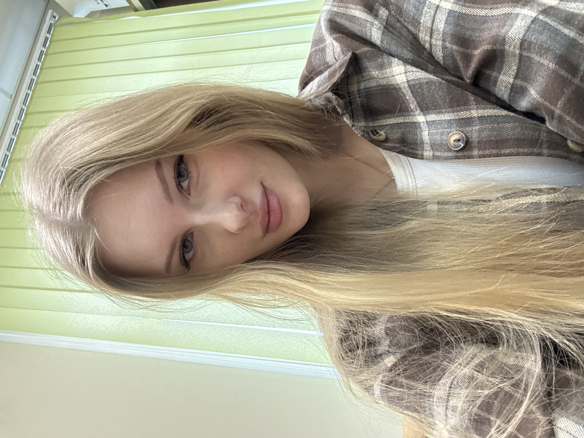 A young woman with long blonde hair is angled toward the camera. She has fair skin and light-colored eyes. She wears a white top and a brown plaid flannel shirt. Behind her are light green vertical blinds. The location is unknown and the city of Perm, Russia cannot be verified from this image.