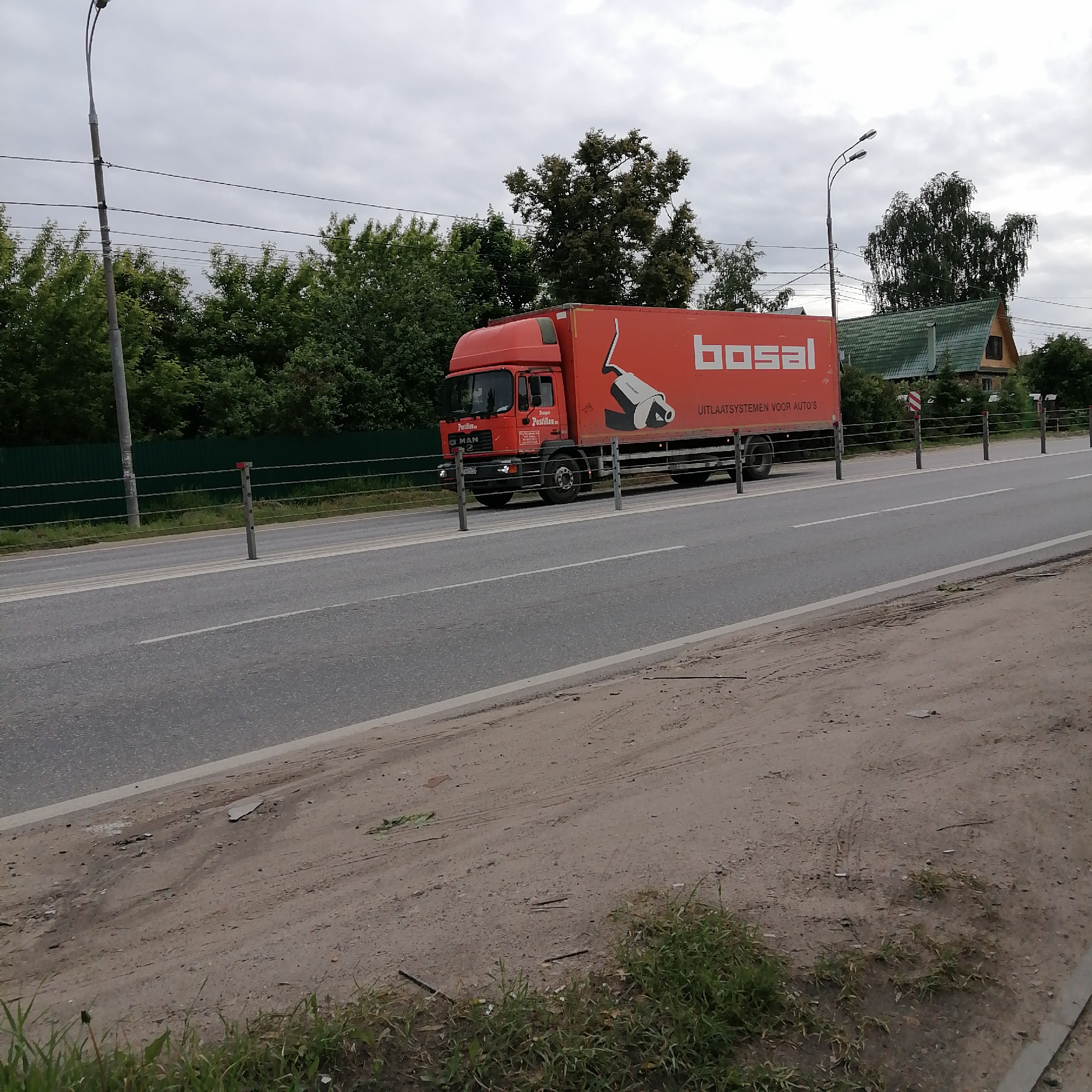 An overcast daytime scene shows a large red semi-truck driving on a two-lane asphalt road. The truck features a red cab and a red box trailer. On the side of the trailer, the brand name "bosal" is prominently displayed in white, along with the text "UITLAATSYSTEMEN VOOR AUTO'S" (Dutch for "EXHAUST SYSTEMS FOR CARS"). An illustration of an exhaust system component is also visible next to the "bosal" logo. On the truck's cab door, text that appears to read "Transport Postillion" and "MAN" is visible.
A wire-rope barrier separates the road from a grassy and dirt shoulder on the right, which then leads to a line of green trees. To the left of the road, another green fence and dense foliage are present. Several dark grey lampposts with twin light fixtures line the road. In the background, behind the trees on the right, part of a house with a green, steeply pitched roof can be seen. The sky is completely cloudy, indicating a dull or overcast day. No people are visible.