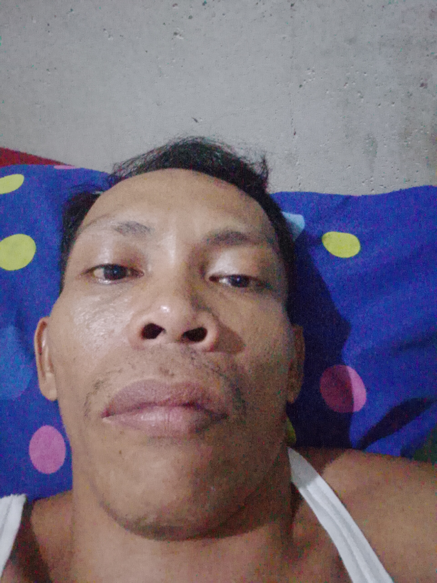 A man is lying down on a pillow with a blue and multi-colored polka dot pattern. He is wearing a white tank top. The background is a gray wall. The location is Bukit Ketok, Indonesia.