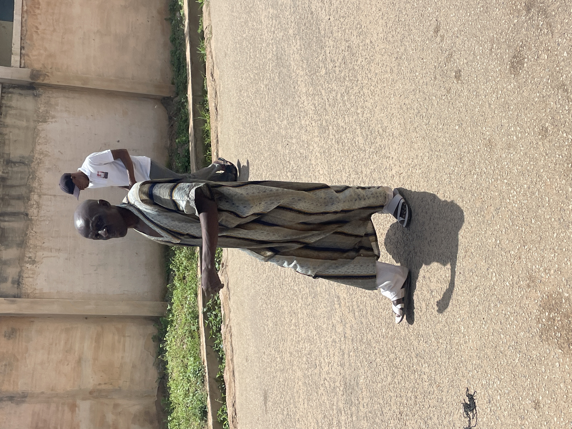 The image depicts a street scene in Jos, Nigeria. In the foreground, a bald man wearing a long, striped kaftan and white trousers is walking on a paved road. He is wearing sandals and his right arm is extended forward, with his index finger pointing. In the background, another person wearing a white polo shirt, grey trousers, and a black baseball cap is walking away from the camera. A low, textured concrete wall runs along the background, with overgrown green vegetation in front of it. The road surface is asphalt with visible aggregate.