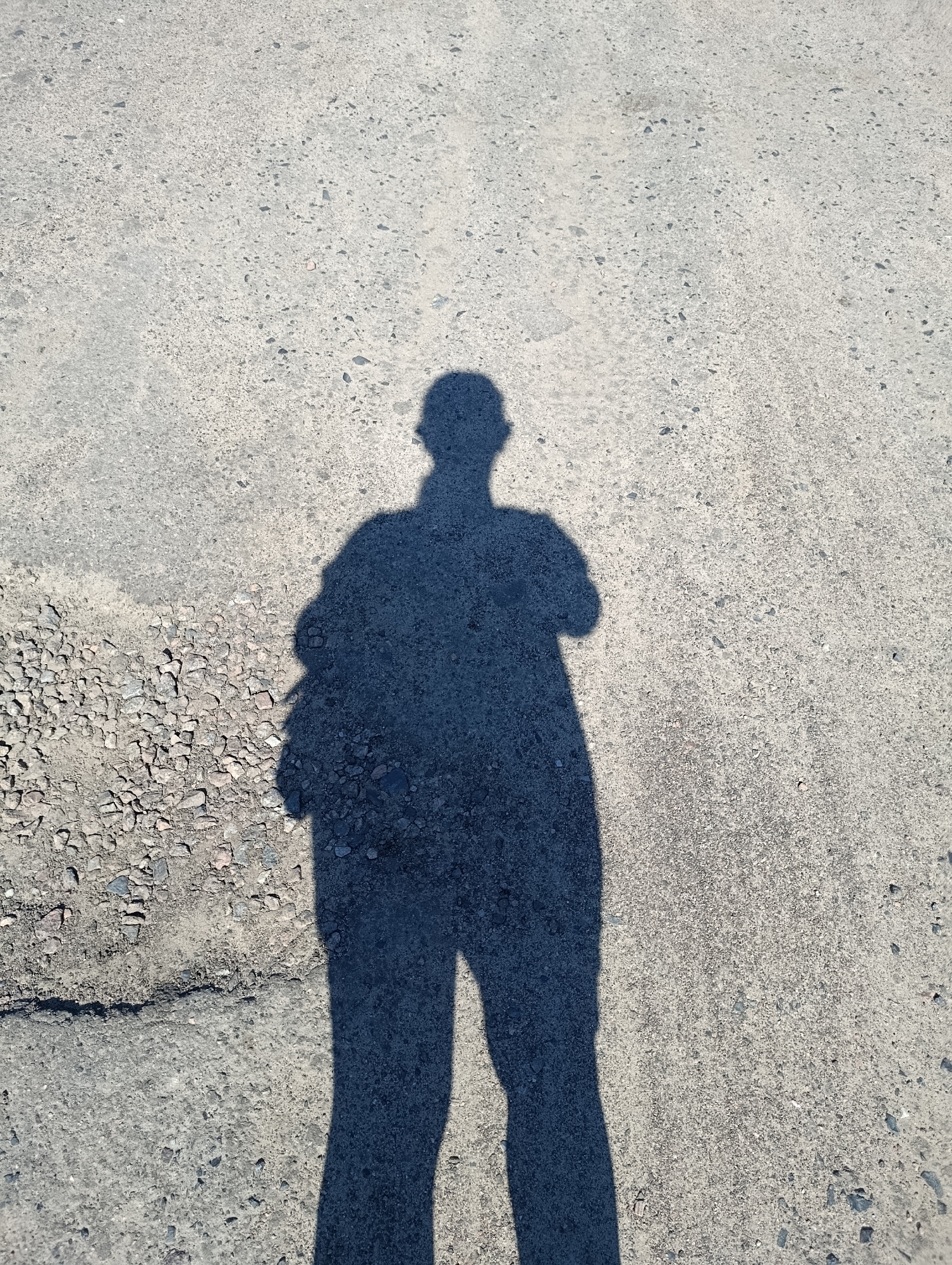The image shows a shadow of a person standing on a textured asphalt surface. The shadow is cast by an unseen light source from above and to the right of the subject, creating a distinct silhouette on the ground. The asphalt appears to be a road or sidewalk, with a rough, gravel-like texture in some areas, particularly towards the bottom left where it transitions to loose stones. The shadow depicts a person with their arms crossed and a bag or backpack slung over one shoulder. The person's head is visible, and they appear to be looking forward. The lighting suggests it is daytime. The image's context is a street in Cherkasy, Ukraine.