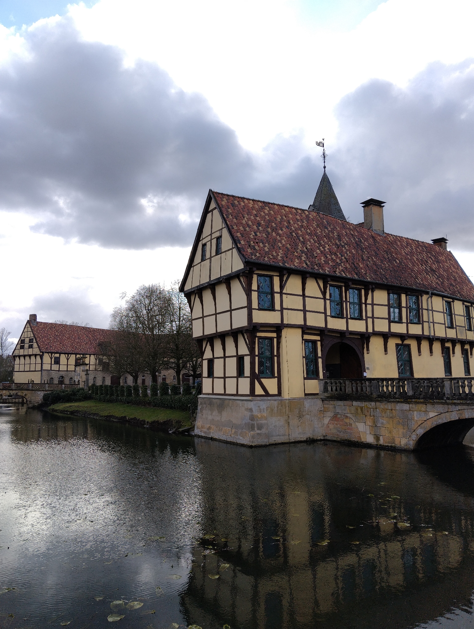 The image depicts a tranquil scene at a moated architectural complex in Burgsteinfurt, Germany, likely Schloss Burgsteinfurt. The primary focus is a large, two-story half-timbered building situated directly on a dark, rippled body of water, which reflects its facade and the overcast sky. This building features light cream-colored infill panels contrasted with dark brown timber framing, a prominent gabled red-tiled roof, and multiple windows with dark frames, many appearing tinted or reflecting blue hues. An arched stone bridge with an ornate balustrade connects the building's ground level to the adjacent land. Behind this structure, a dark grey conical spire topped with a weathervane is visible against the cloudy sky, alongside a stone chimney. To the left and further in the background, a second, similar half-timbered building with a red-tiled roof is present, also bordering the water. A narrow strip of green grass and manicured hedges line the water's edge between the two buildings, backed by several deciduous trees with bare branches. The sky is largely overcast with significant grey cloud cover and some brighter, lighter cloud formations. No individuals or discernible actions are present in the frame. The water surface contains scattered circular lily pad-like vegetation.