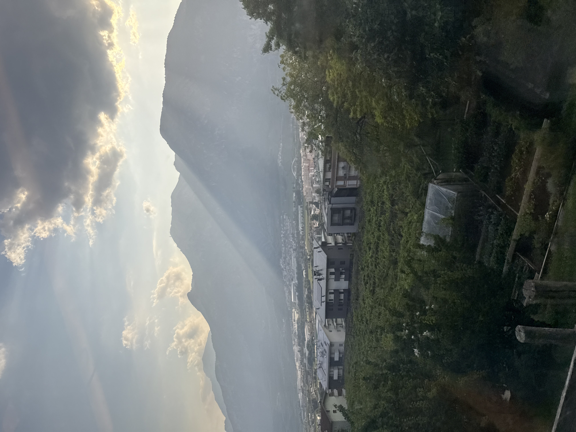 The image shows a panoramic view of the city of Trento, Italy, as seen from a higher vantage point, likely a hillside overlooking the city. The foreground features a lush green vineyard with rows of vines stretching across the landscape. Several modern residential buildings with flat roofs, some equipped with solar panels, are situated on a gentle slope above the vineyard. Further in the midground, a sprawling urban area of Trento is visible, characterized by a mix of buildings, including some taller structures and what appears to be a stadium or large dome-like structure in the distance. The background is dominated by imposing, mist-shrouded mountains, their peaks softened by atmospheric haze. Dramatic sunbeams pierce through the clouds, casting a striking light and shadow pattern across the mountains and the valley. The overall impression is one of a serene, sun-drenched afternoon in an Italian city nestled amongst dramatic natural scenery.