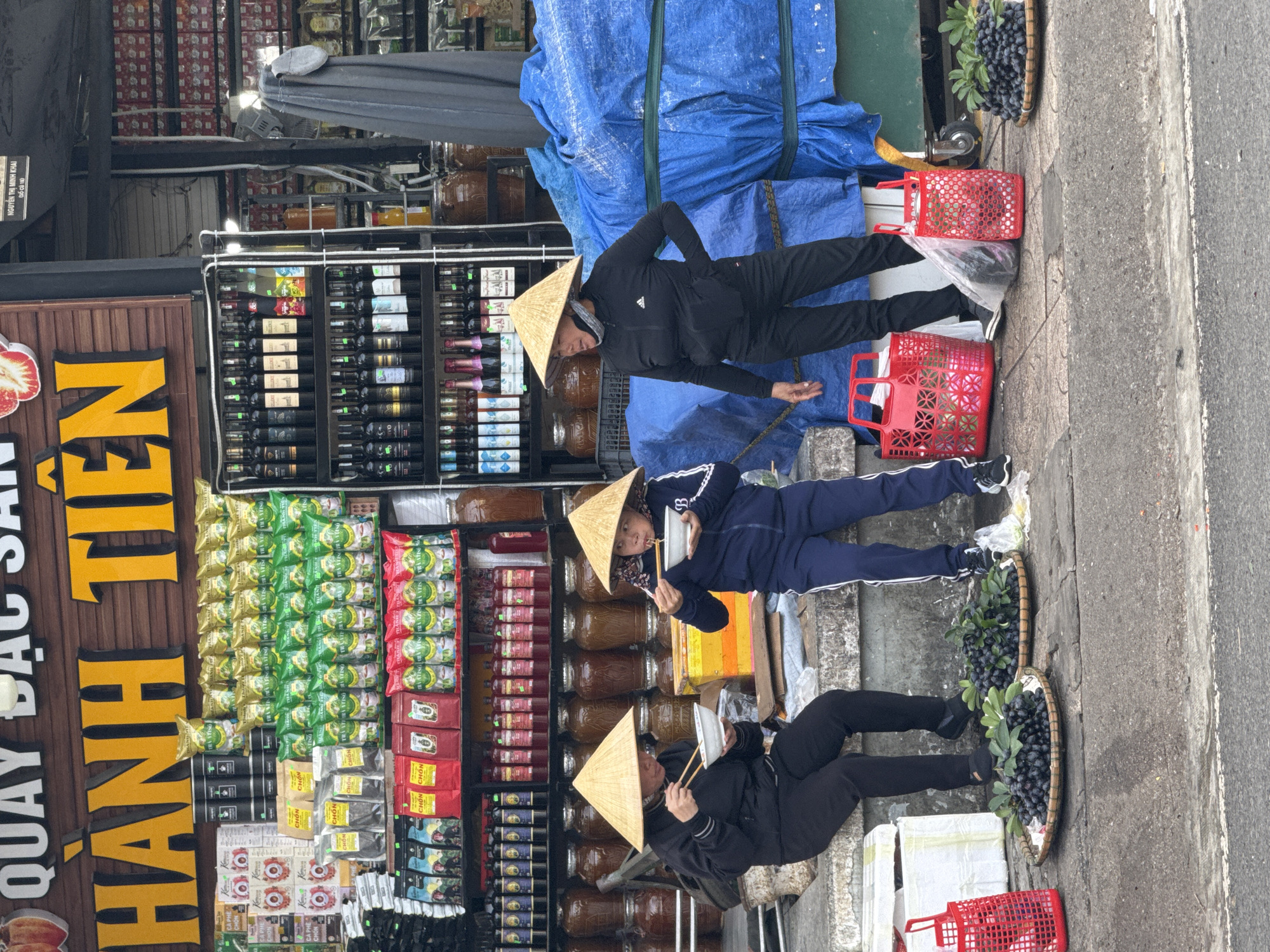 Three people wearing traditional Vietnamese conical hats are sitting on the pavement in front of a shop. They are eating from bowls with chopsticks. The shop displays a variety of goods, including packaged teas, coffees, and bottled drinks. Blue-covered bundles are stacked behind them. Two baskets filled with a dark fruit are placed on the pavement. The shop's sign reads "QUAY ĐẠC SẢN HẠNH TIÊN." Another smaller sign includes the text "NGUYỄN THỊ MINH KHAI," along with "(SỐ CŨ 18)," which can be interpreted as a previous address. Red plastic baskets are also near the seated individuals. The location appears to be a commercial area, with a variety of goods for sale, which indicates Da Lat, Vietnam.