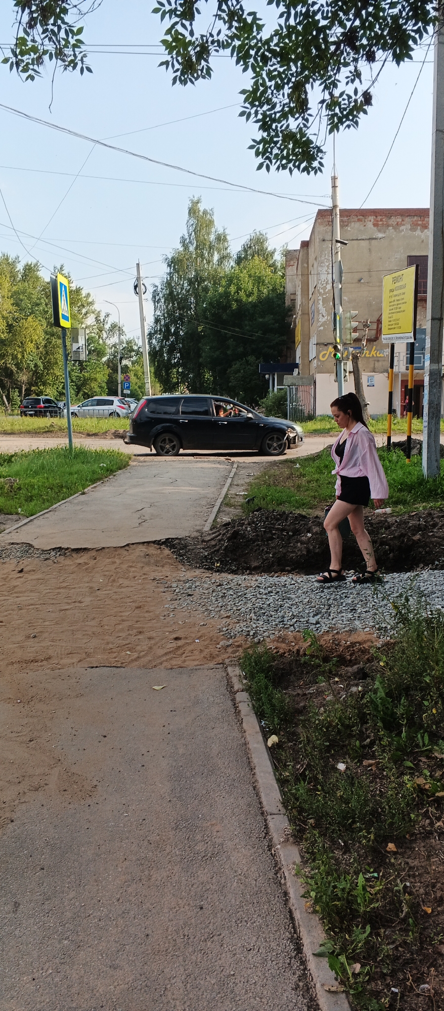 The image depicts a street scene in Izhevsk, Russia. A young woman with dark hair, wearing a pink shirt and black shorts, walks on a gravel path next to a construction zone. To her left, a dark-colored station wagon is parked on the road. A pedestrian crossing sign is visible in the background, along with trees and buildings. The overall environment suggests an urban setting with ongoing roadwork.