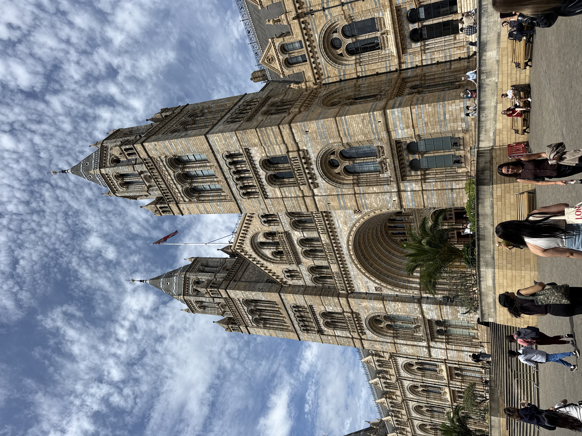 The image depicts the Natural History Museum in London, United Kingdom, on a sunny day with a sky filled with scattered clouds. The museum's facade is prominently featured, showcasing its intricate architectural details, including arched windows, towers, and decorative stonework. A British flag flies atop one of the towers. People are present in the foreground, walking and sitting on benches, suggesting the museum is open to visitors. A variety of clothing is worn by the people in the image. The ground appears to be paved with light-colored stone.
