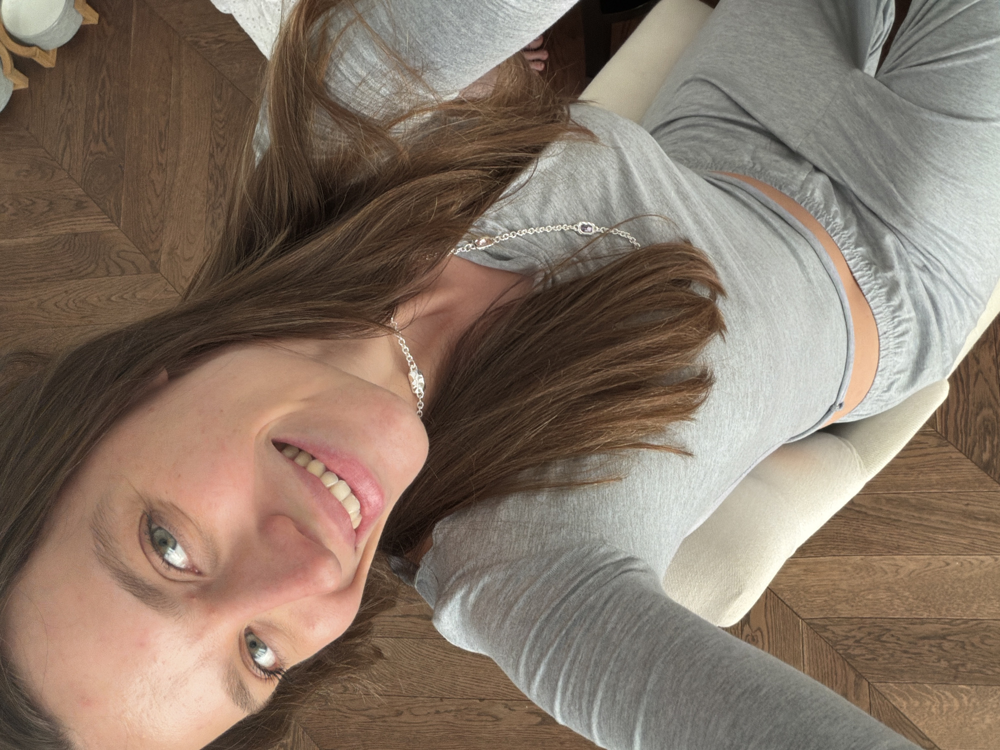 A woman is lying on a white upholstered chair in a room with a herringbone wood floor. She is wearing a light grey long-sleeved shirt and matching pants, a silver necklace, and is smiling at the camera. A glimpse of her skin can be seen between the shirt and pants. Part of a candle on a wooden stand can be seen in the upper-left corner of the image. The location cannot be determined from the image.