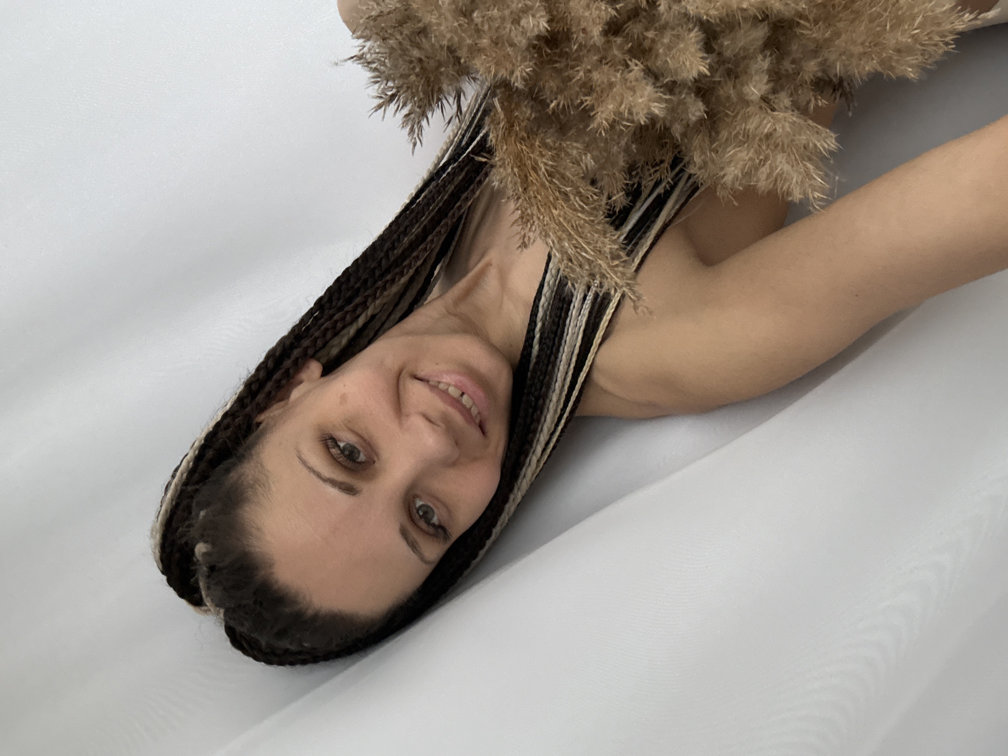 A person with braided hair is lying on a white surface, holding a bunch of dried plants in front of them. The location is not specified in the prompt, only Rusanovo, Russia is.