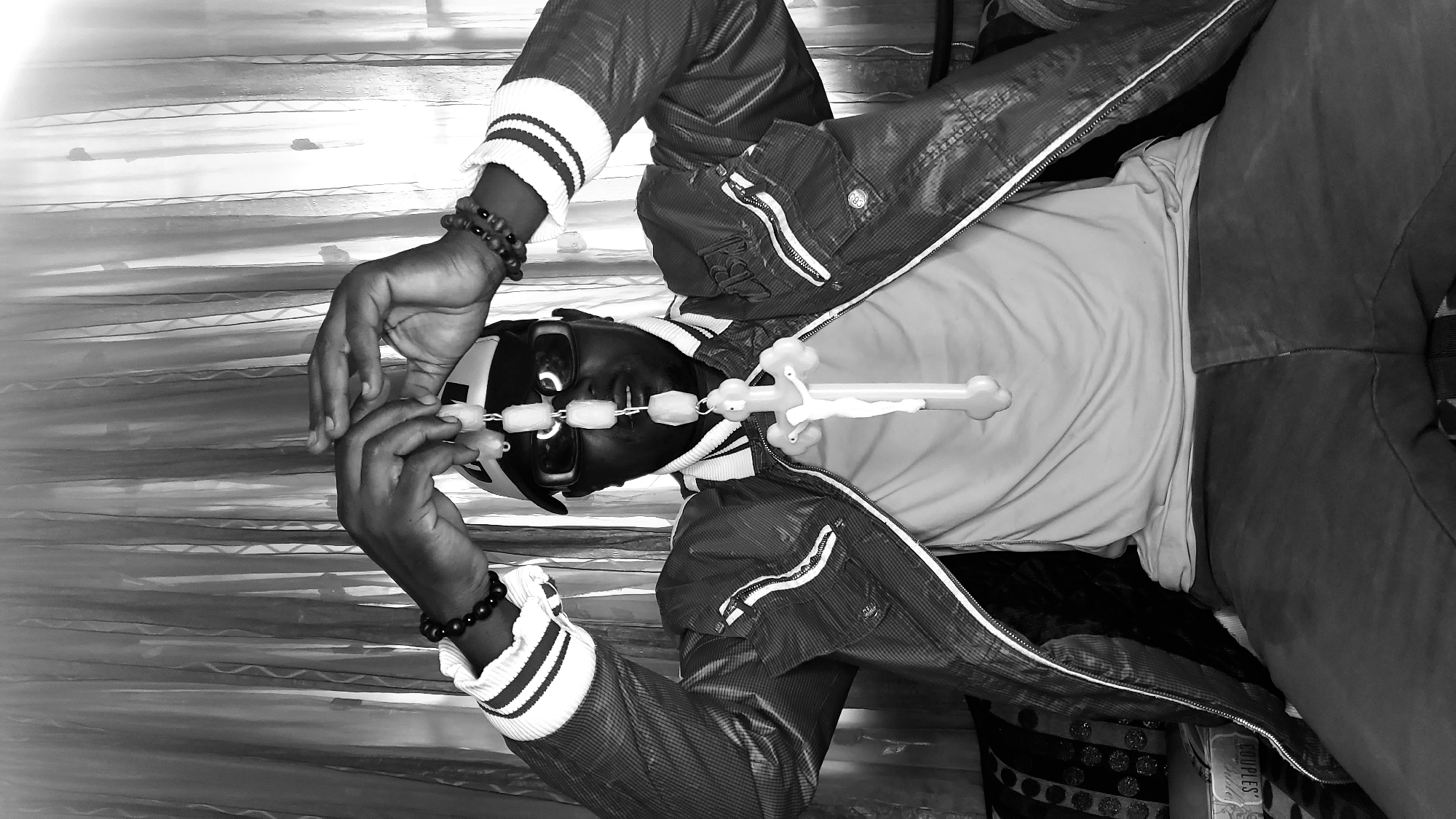 A black and white, waist-up, medium shot shows a Black male sitting, wearing a baseball cap, sunglasses, a light-colored t-shirt, and a patterned jacket with white striped cuffs. He holds a string of large, light-colored beads with a cross at the end, directly in front of his face, with both hands clasped above it. The background consists of vertically draped, sheer curtains. The overall scene is indoors, likely in a residential setting within Sabon Tasha, Nigeria.