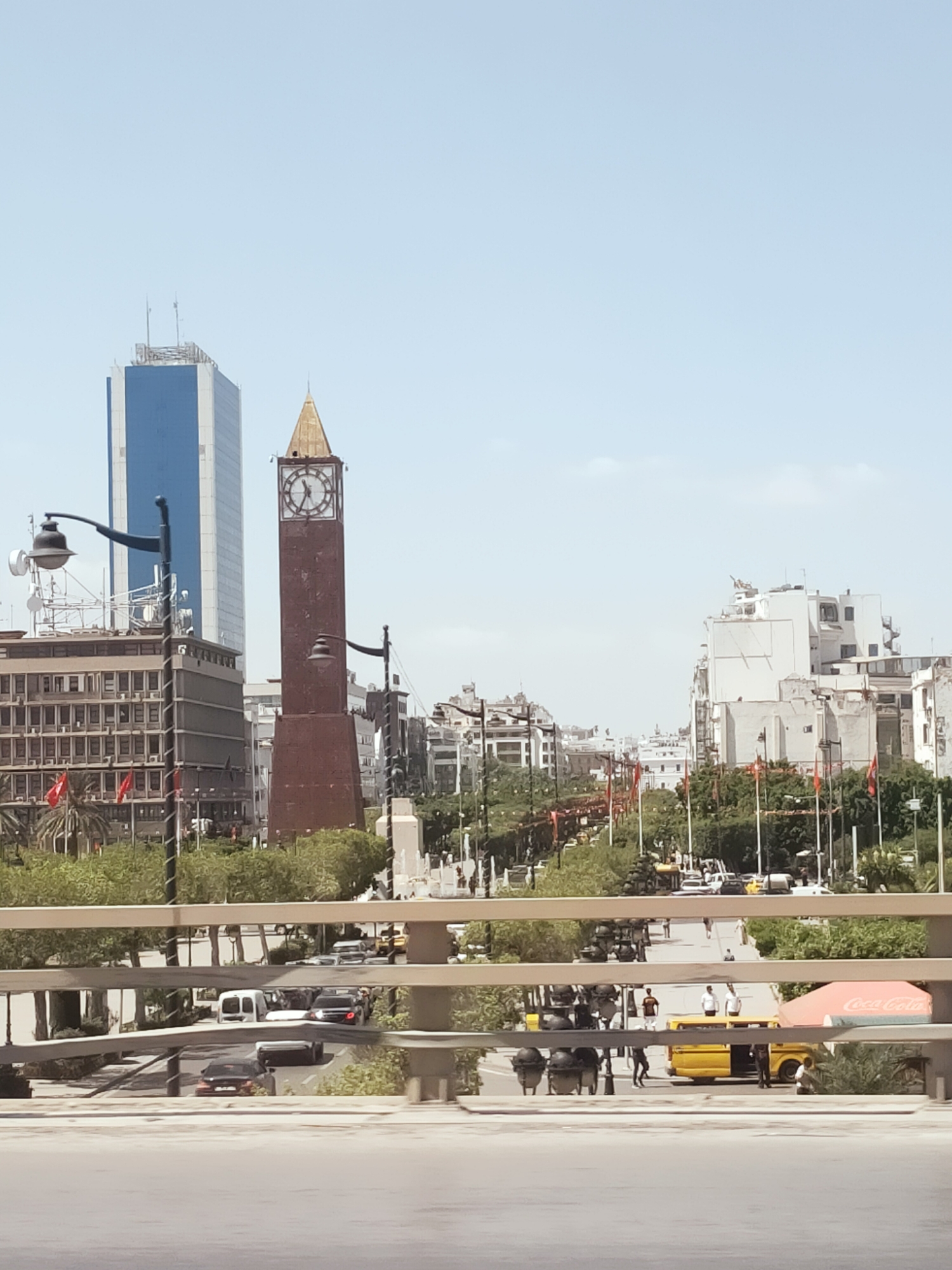 The image shows a street scene in Tunis, Tunisia, on a clear day. In the background, a tall blue and white skyscraper stands next to a prominent clock tower with a golden spire. The clock tower appears to be made of weathered, reddish-brown material. Several other buildings, some modern and some older, are visible along the street, which is lined with trees and flags. Cars, including a white van and a yellow bus, are driving on the street. People are walking on sidewalks and crossing the street. In the foreground, a metal railing suggests the photo was taken from an elevated position, such as a bridge or overpass. A red and white Coca-Cola umbrella is visible on the right side of the street.