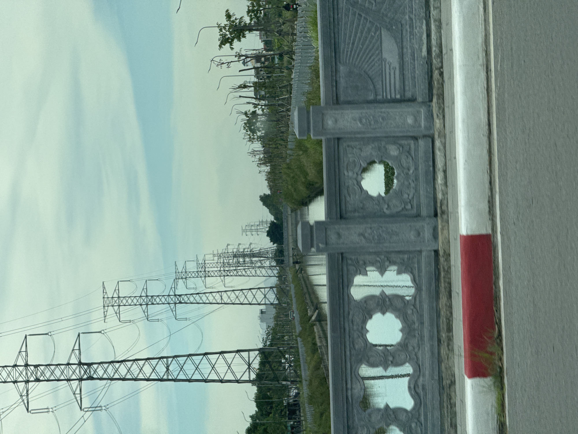The image depicts a scene in Hoa Lư Ward, Vietnam, featuring a canal bordered by vegetation and a row of electricity pylons extending into the distance. In the foreground, a concrete bridge railing with decorative cutouts and reliefs overlooks the canal. A red and white striped barrier runs along the edge of the roadway adjacent to the bridge. The sky is overcast.