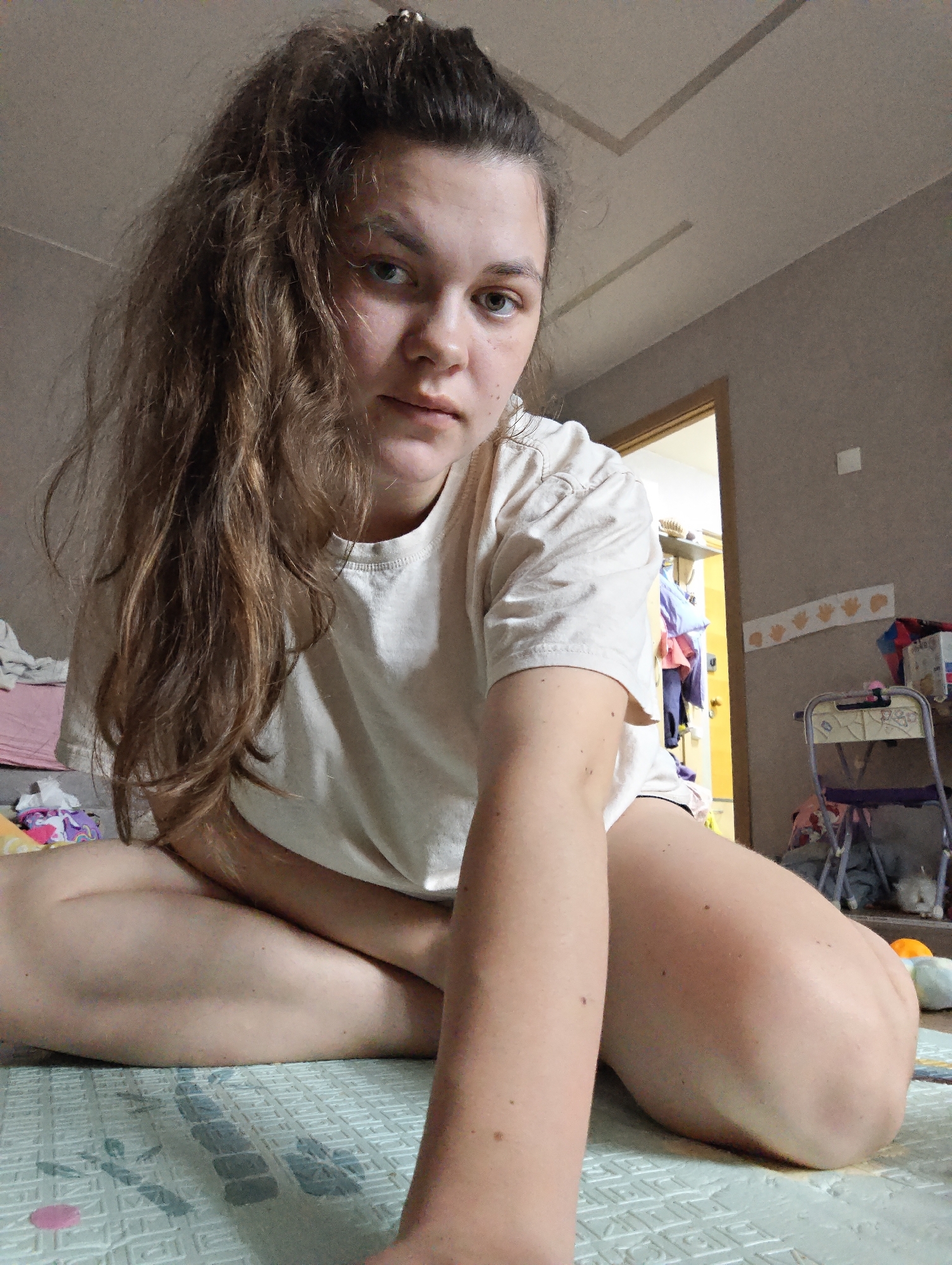 A young adult female is depicted seated cross-legged on a light-colored, patterned foam mat in an indoor residential setting located in Chaikovsky, Russia. She wears a light-colored short-sleeved t-shirt and has long, wavy brown hair, partially secured in a high ponytail. Her eyes are light, and she looks directly towards the camera with a neutral expression, her left arm extended forward and partially cropped. The room features light grey-brown walls and a light-colored ceiling with decorative linear indentations. In the background, an open doorway with a wooden frame reveals a brightly lit adjacent room with yellow walls and hanging garments. To the subject's left, crumpled clothing items rest on a pink surface, alongside a pack of patterned tissues. To her right, a small, light-colored chair with purple components is visible, partially obscured by various items, including a white plush toy. Above the chair, cut-out paper handprints are affixed horizontally to the wall, and a white square light switch or outlet is also present on the wall.