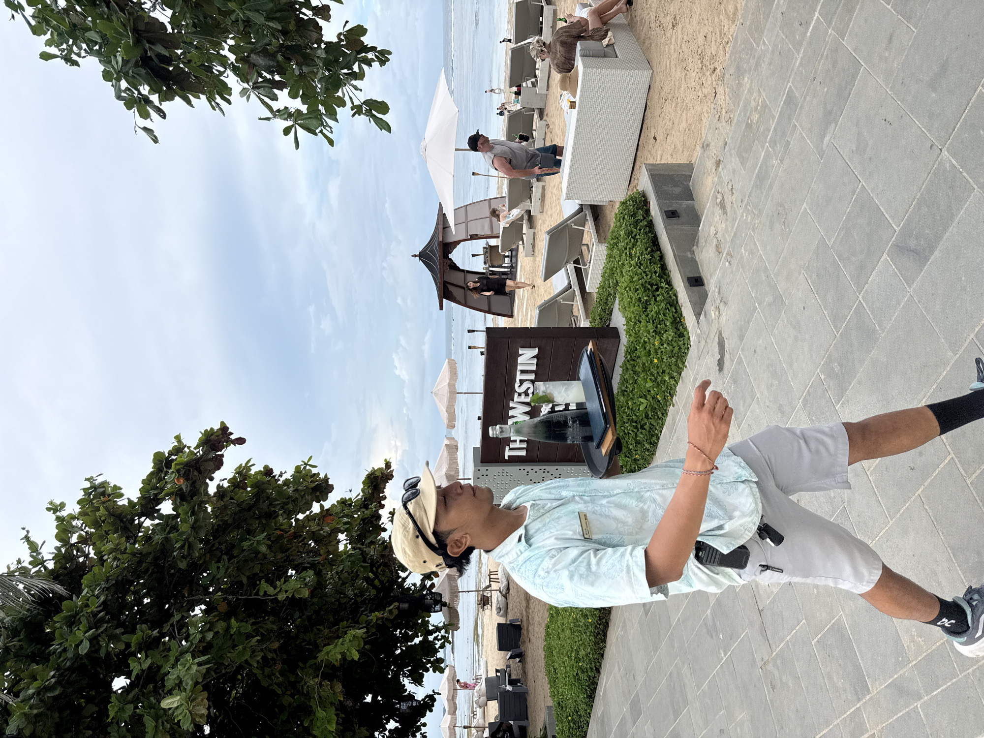 A photo of a beachside setting in Nusa Dua, Indonesia shows a man in the foreground, wearing a light blue patterned shirt, khaki shorts, a tan baseball cap, and dark socks with sneakers. He stands on a paved walkway, holding his left hand up as if gesturing. Behind him is a sign for "The Westin," indicating the location of the hotel. In the background is a beach with white umbrellas, lounge chairs, and a small gazebo. There are several people lounging on the beach. The sky is partly cloudy, and a tree is visible on the left side of the image.