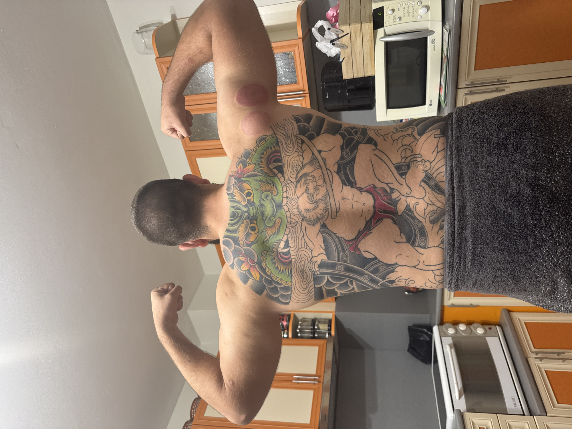 A man is depicted from behind, flexing his arm muscles, in what appears to be a home kitchen or an open-plan living area in Stara Zagora, Bulgaria. He is shirtless, with a dark grey towel wrapped around his lower body. His back is extensively covered by an elaborate, unfinished Irezumi-style tattoo. The tattoo features a vibrant green dragon or mythological creature on his right side and a human-like warrior figure on his left side, with elements colored in green, red, black, and yellow, while other parts remain in outline.
On his left upper arm, two distinct circular red marks from cupping therapy are visible. The man has short, dark hair and a beard.
The background shows various kitchen elements, including light beige and darker brown framed cabinets, some with glass inserts, and others with solid orange-brown panels. A white LG "WaveDOM" microwave oven sits on a dark countertop, alongside another white appliance with "VELCO" branding and three knobs on top, possibly a small oven or toaster. Other items like a wooden chopping board, plastic bags, and a glass container are also visible. The walls are light-colored. The scene captures the man posing, showcasing his physique and tattoo, likely after a recent cupping therapy session.