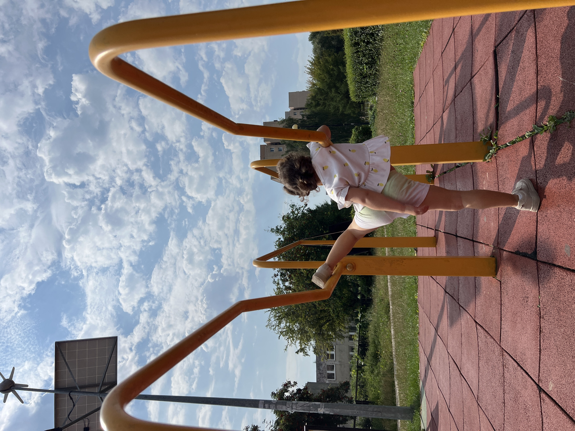 A young child with curly brown hair is performing a gymnastic pose on an outdoor fitness apparatus. The child is wearing a light-colored shirt with a pattern of yellow lemons and light green shorts, along with white sneakers. The child's right leg is extended upwards and backwards, while their left leg is bent and supporting their weight as they grasp a yellow horizontal bar. The fitness equipment is made of bright yellow metal and is situated on a red rubberized play surface.
In the background, there is a grassy area with trees and bushes. Beyond the greenery, several apartment buildings are visible. The sky is blue with scattered white clouds. A solar panel and a weather vane are mounted on a pole to the left of the frame. The scene appears to be an outdoor public space, likely a park or playground, in the city of Lublin, Poland.