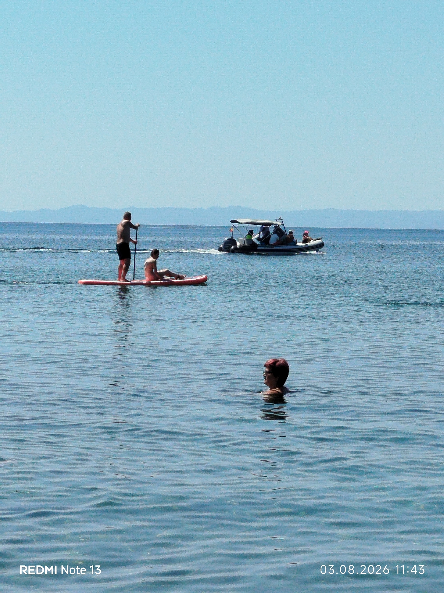 A medium shot captures a clear, calm sea under a bright blue sky. In the foreground, a person with short brown hair is partially submerged in the water, looking to their left with their head and shoulders visible. In the mid-ground, a red paddleboard floats on the water. A shirtless man stands on the paddleboard, holding a paddle. A second person, also partially visible, sits on the paddleboard in front of the man. Further in the distance, a speed boat with a canopy is cruising, carrying several passengers. The background shows a hazy, mountainous coastline. The image has a timestamp in the bottom right corner: "03.08.2026 11:43" and a text overlay in the bottom left corner: "REDMI Note 13". The scene appears to be a leisure activity taking place in the waters of Sithonia, Greece.