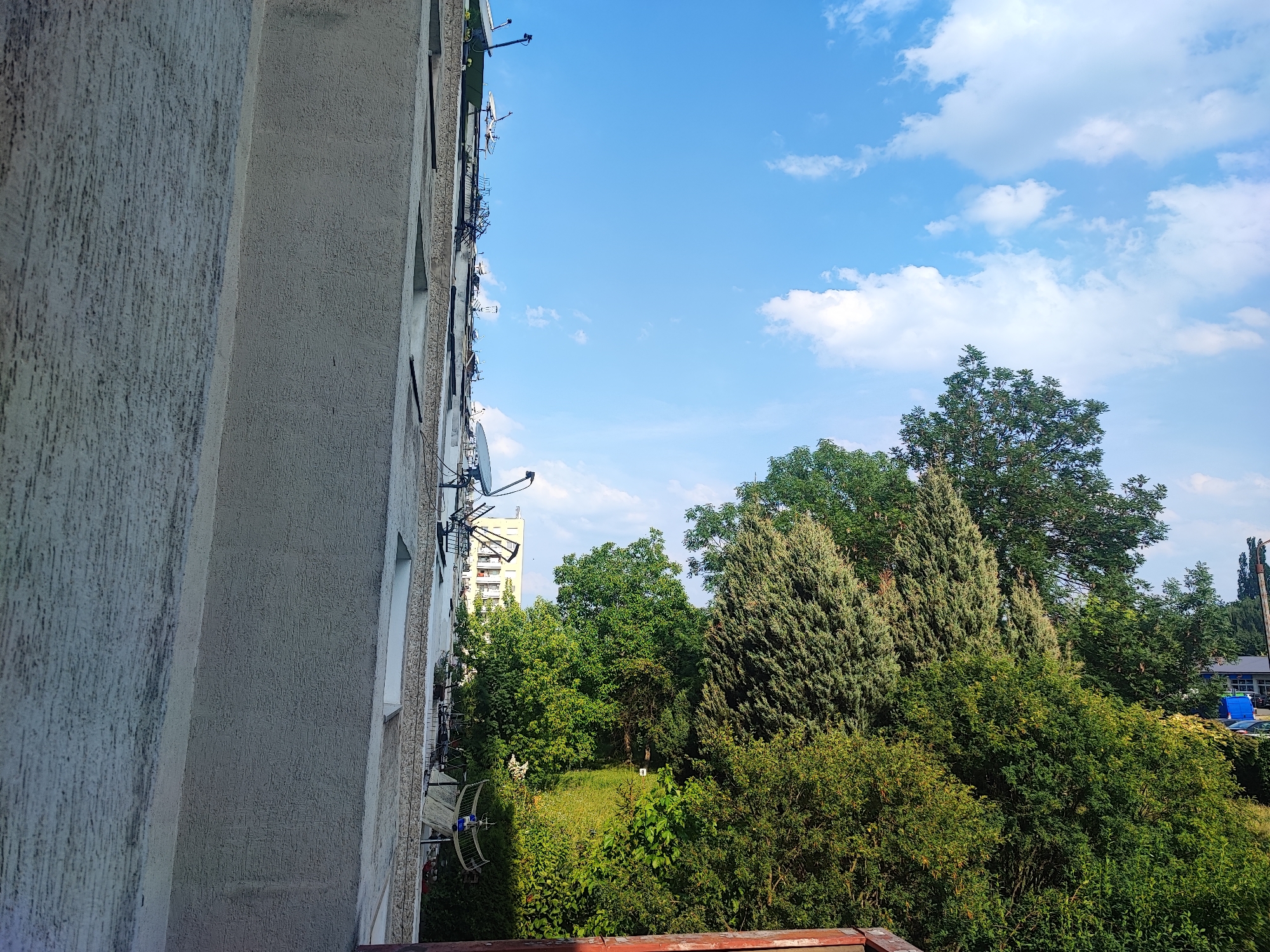 A vertical medium shot captures the exterior of a concrete building and a lush green park under a bright blue sky dotted with fluffy white clouds. The building, on the left, is textured with a rough, light-colored stucco and features multiple satellite dishes mounted on its side. Windows are visible, some with dark frames. The sky occupies the upper half of the image, suggesting daytime.
To the right of the building, the park is a dense expanse of various trees and shrubs, ranging in color from deep green to lighter, almost yellowish-green foliage. Prominent are several tall, conical evergreen trees, possibly cypresses or junipers, interspersed with broader deciduous trees. The park appears well-maintained, with grassy areas visible beneath the trees. In the distance, a tall apartment building can be seen peeking through the trees, and a hint of a road with cars and a building with a blue roof suggests a semi-urban or suburban setting.
The image composition, with the building dominating the left side and the park extending into the right and background, creates a sense of contrast between urban infrastructure and nature. The lighting suggests it is a sunny day, possibly late morning or early afternoon, given the clear sky and the shadows cast by the trees. No people or specific activities are depicted. The overall mood is serene and bright, highlighting a pleasant outdoor environment. There is no visible text in the image.