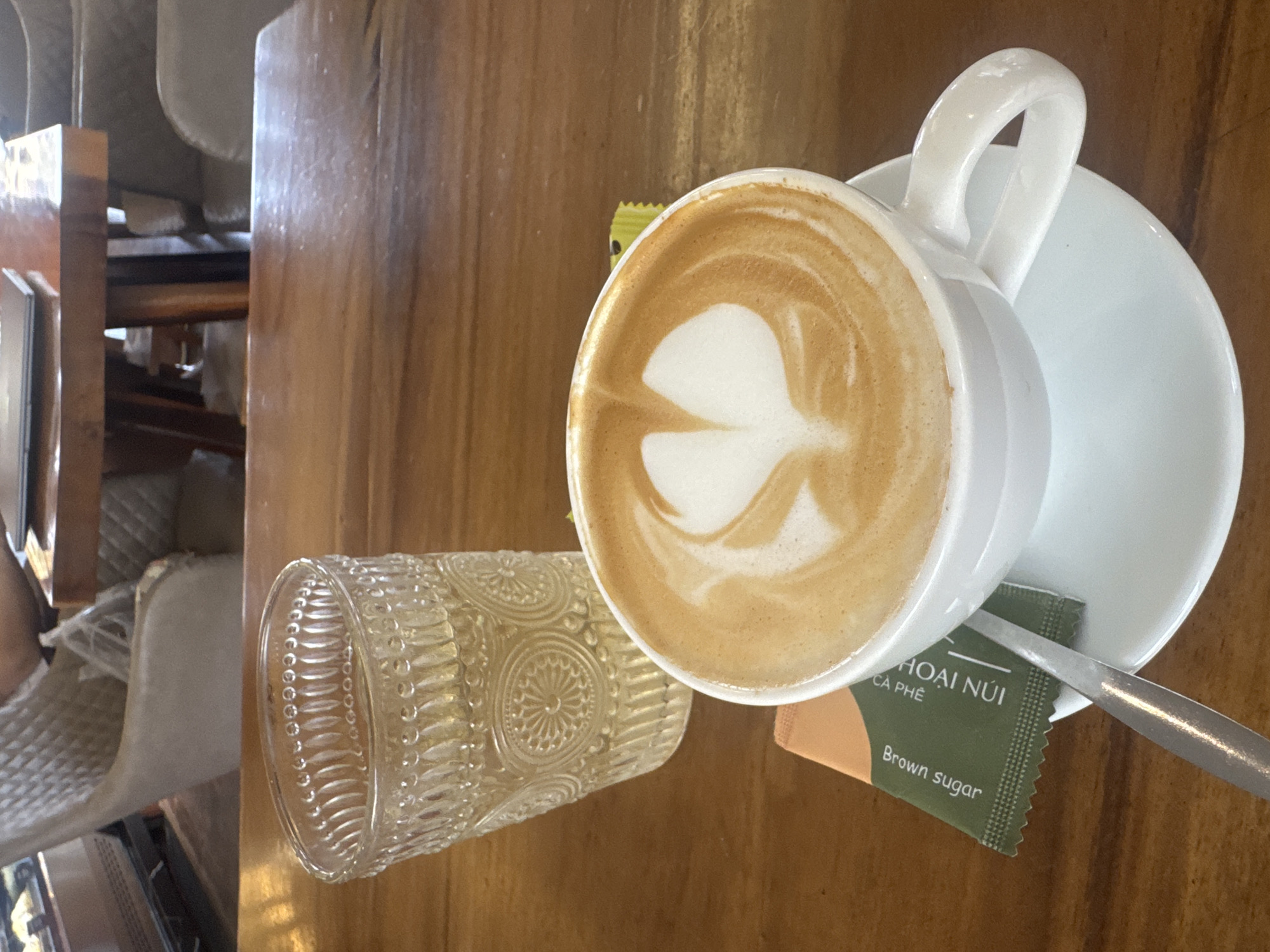 In a cafe in Bac Nha Trang Ward, Vietnam, a white ceramic cup of coffee with latte art sits on a saucer. Next to it is a glass of water and a packet of brown sugar, with a spoon resting on the saucer. The cafe's interior, featuring wooden tables and upholstered chairs, is visible in the background.