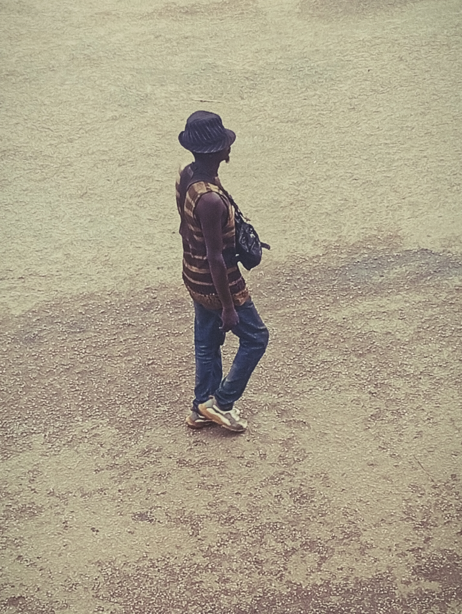 A person, viewed from above and to the side, walks across a dusty, gravelly ground. They are wearing a dark bucket hat, a sleeveless shirt with horizontal brown and yellow stripes, and blue jeans. A dark bag is slung across their chest. The person is also wearing athletic shoes with light and dark accents. The scene is outdoors, with a textured ground that suggests it could be a road, pathway, or open area. The lighting appears even, suggesting it might be daytime, possibly overcast. There are no visible signs of text in the image. The overall impression is of a person casually moving through an open space.