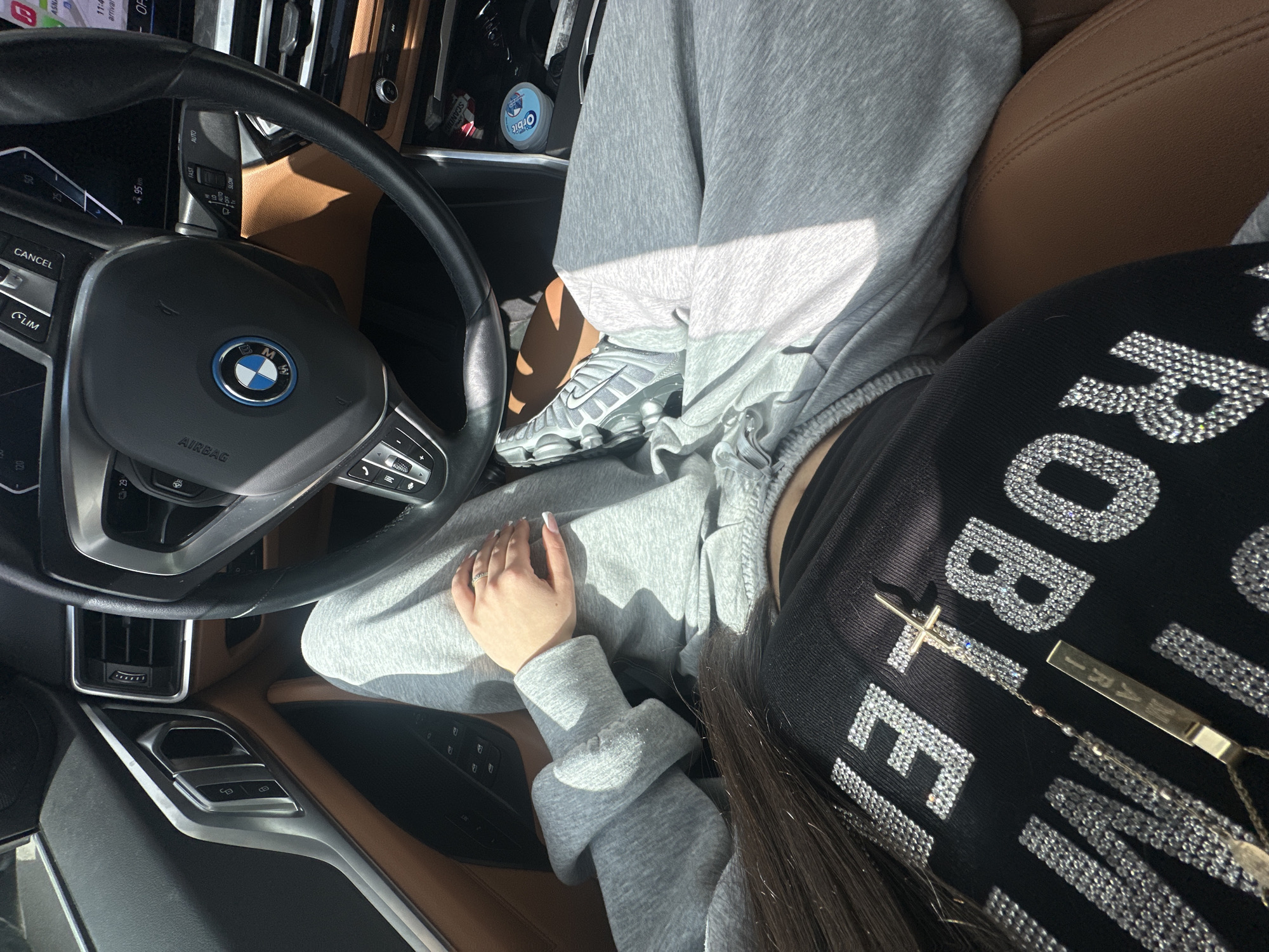 A first-person, interior car shot shows a person sitting in the driver's seat of a BMW with a brown leather interior. The person is wearing gray sweatpants and a black shirt with the word "ROBLE" written in rhinestone letters. A pair of gray sneakers is visible. A cross necklace hangs from the person's neck. The car's dashboard, steering wheel, and central console are visible. A package of Orbit gum is placed in the pocket of the driver's seat. The rearview camera shows an unidentified street location in Yerevan, Armenia. The screen of the rearview camera displays a street named "ASN" and the destination ETA as 11:41.