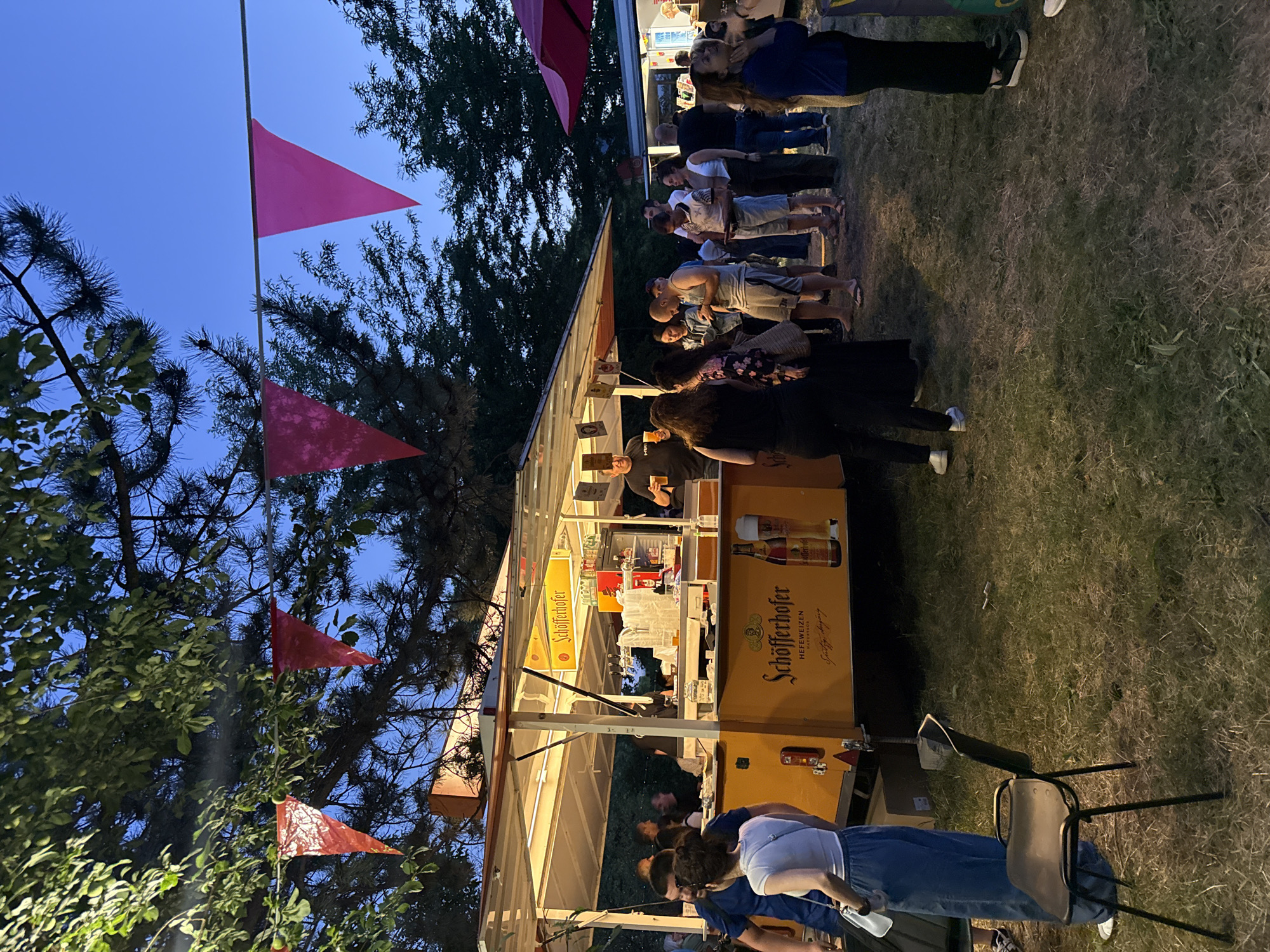 The image depicts an outdoor gathering at dusk, likely an event or festival. In the foreground, a yellow food truck with a "Schöfferhofer" logo is illuminated by interior lights, serving drinks. Several people are standing in line or conversing around the truck. Red and pink triangular flags are strung across tree branches overhead, adding a festive atmosphere. The ground is covered with dry grass and dirt. In the background, more people and other structures, possibly more food vendors or tents, are visible. The sky is a deep blue, indicating the late hour. The setting appears to be a park or open field. No specific landmarks of Stara Zagora, Bulgaria, are discernible in the image.