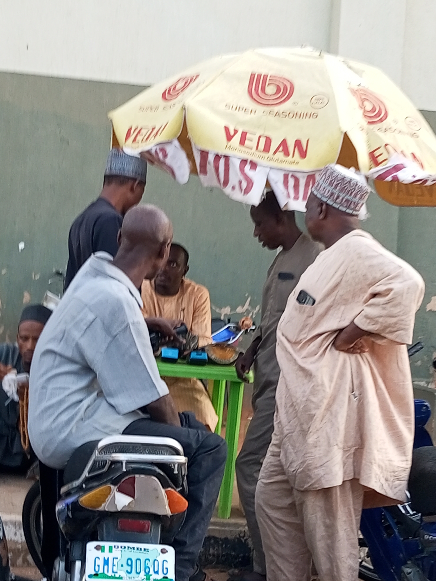 In Gombe, Nigeria, a group of men are gathered under a yellow umbrella advertising "VEDAN SUPER SEASONING." One man, wearing a light grey shirt and dark trousers, sits on a motorcycle, facing away from the camera. Another man in a beige robe stands to his right, with a mobile phone in his breast pocket. To the left of the motorcycle rider, a man in a dark shirt and cap stands, and further left, another man in a dark cap and dark clothing is visible. In the background, a man in an orange outfit sits behind a green table with items on it, including what appears to be a small electronic device. The overall scene depicts a casual gathering on an outdoor surface, likely a street or market area, given the presence of a motorcycle and street vendors' umbrellas.