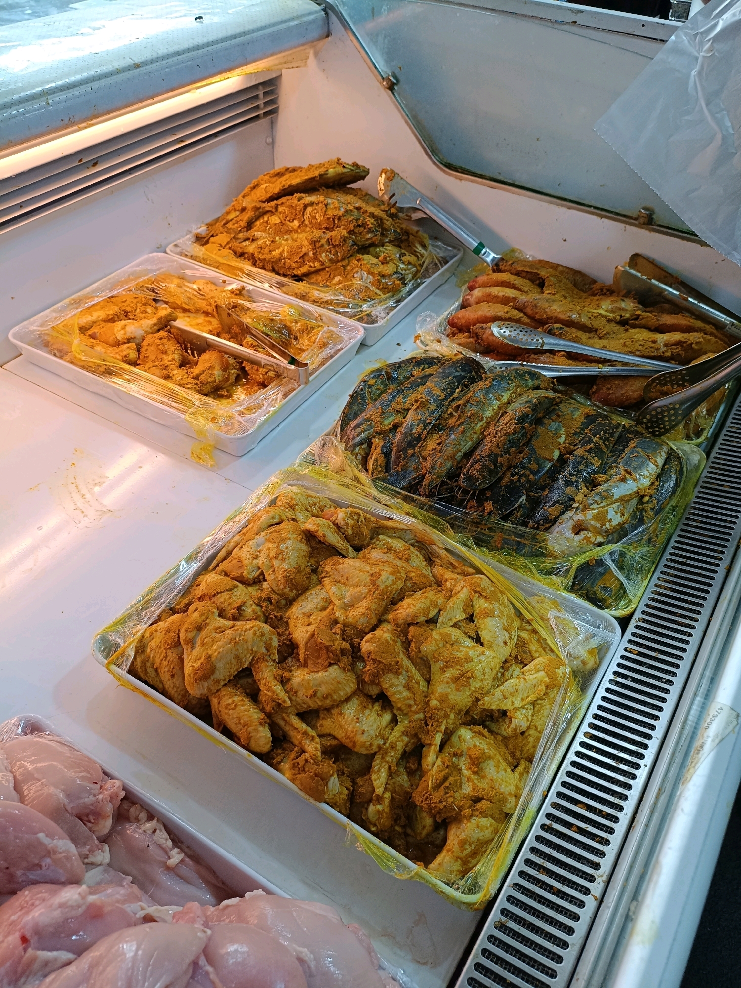 This is a close-up, eye-level shot of a display case in a market, likely in the city of Yogyakarta, Indonesia. The display case is illuminated from above, casting a bright light on the food items. The case contains several trays of raw, seasoned meats and seafood, all wrapped in plastic.
In the foreground, a tray holds an abundance of raw chicken wings, coated in a yellowish spice mixture. To the left, another tray displays more raw chicken pieces, some appearing to be breast meat, also lightly seasoned.
Behind the chicken, a large tray is filled with what appear to be large, dark-colored fish or possibly large shrimp, also seasoned with the same spice blend. Tongs are resting on this tray. Further back, more trays of seasoned food items are visible, including what looks like more fish and perhaps some smaller pieces of seasoned meat.
The overall impression is of a well-stocked display of ingredients ready for preparation, typical of a butcher or fish market. The clean, white surfaces of the trays and the polished metal of the display case suggest a focus on hygiene. The presence of tongs indicates that customers can select their items.