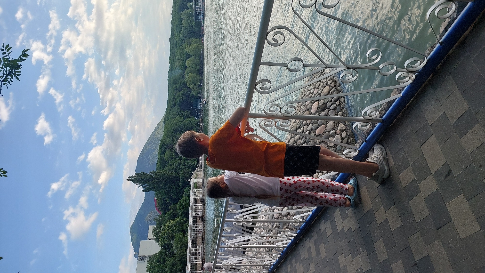 Two children, a boy and a girl, stand side-by-side, leaning on a white metal railing. The boy, wearing an orange t-shirt and black shorts, is on the right. The girl, in a white t-shirt and pink pants with a heart pattern, is on the left. They are facing away from the camera, looking out at a large body of water. Behind the water, a lush green forest covers rolling hills and mountains under a sky filled with scattered white clouds. In the distance, a building with a white facade and a red roof is visible amidst the trees. The scene appears to be a waterfront promenade, likely in the city of Zheleznovodsk, Russia, known for its scenic lakes and surrounding natural beauty. The ground beneath their feet is paved with interlocking gray and black tiles. The railing they are leaning on features decorative scrollwork. The water is calm, reflecting the sky and the surrounding landscape.