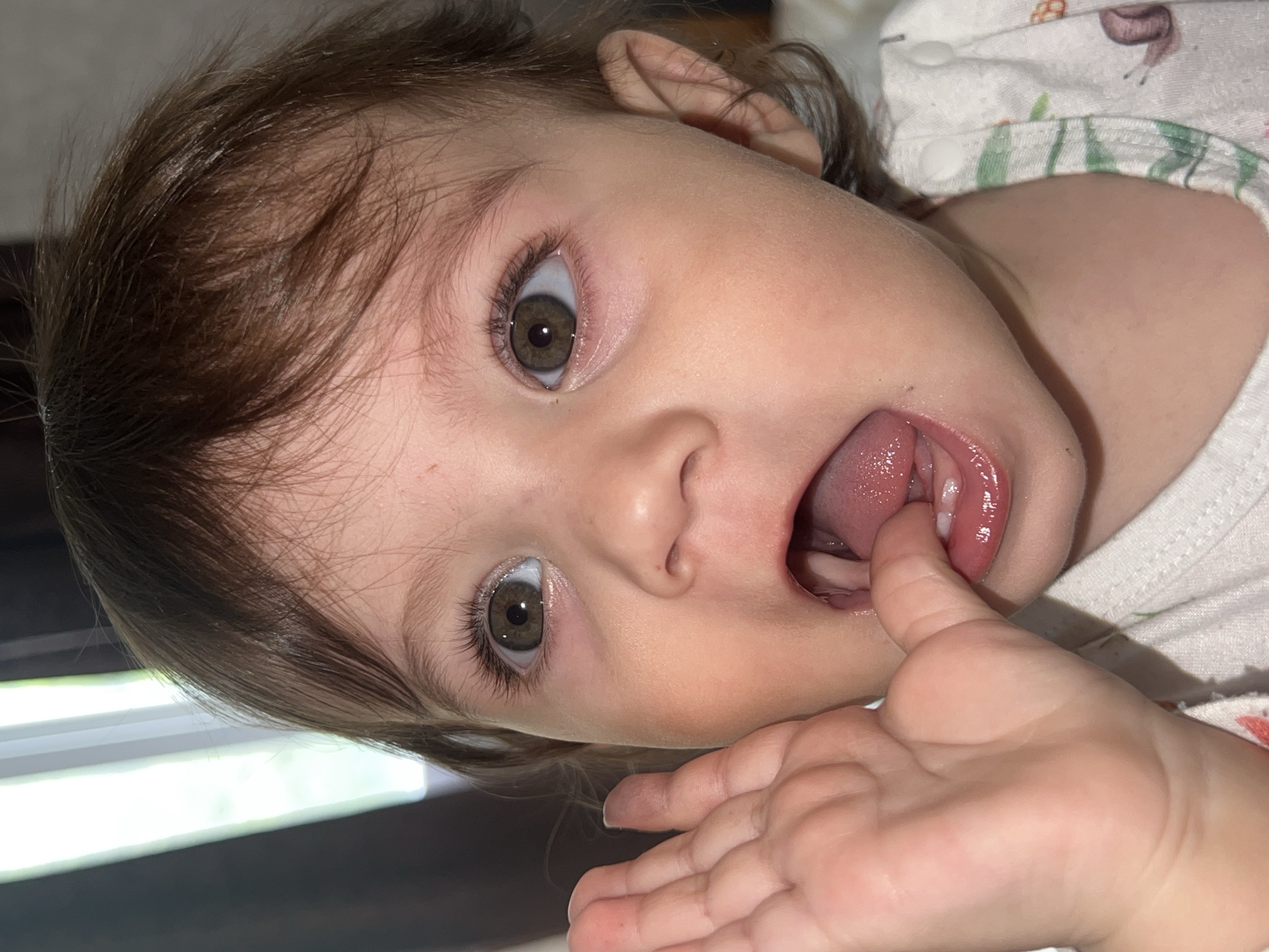 A close-up, rotated image depicts a young child with brown hair and hazel eyes. The child has their mouth open, with a finger inserted between their lips and tongue. The child is wearing a light-colored patterned shirt. The background is out of focus, but appears to be an indoor setting with a light source visible. There is no information to indicate a location in the city of Elena, Bulgaria.