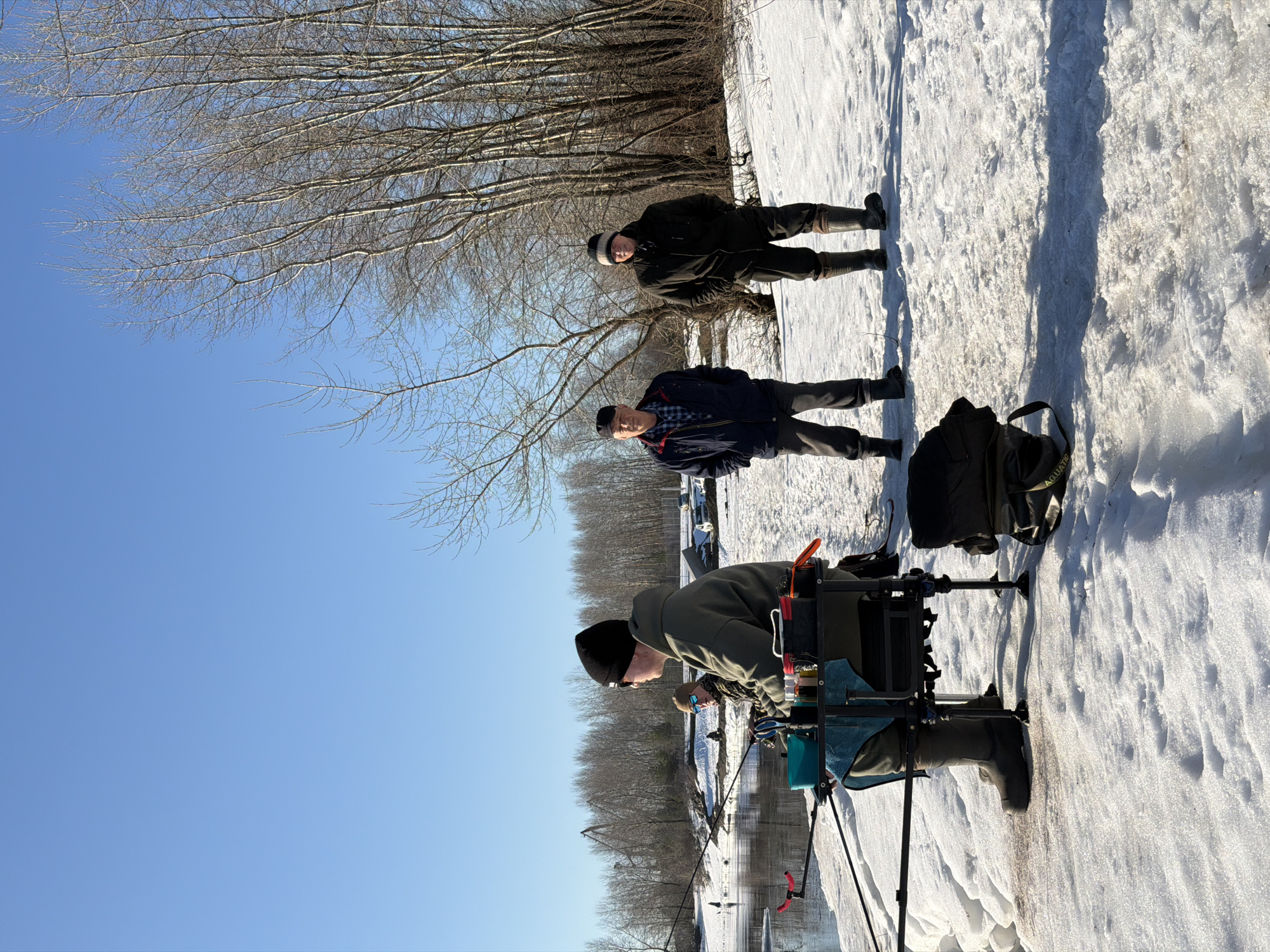 Three men are ice fishing on a snow-covered area. One man is seated, tending to fishing equipment. Two men stand behind him. The environment includes trees without leaves and a clear blue sky. The location is Noviy, Russia.