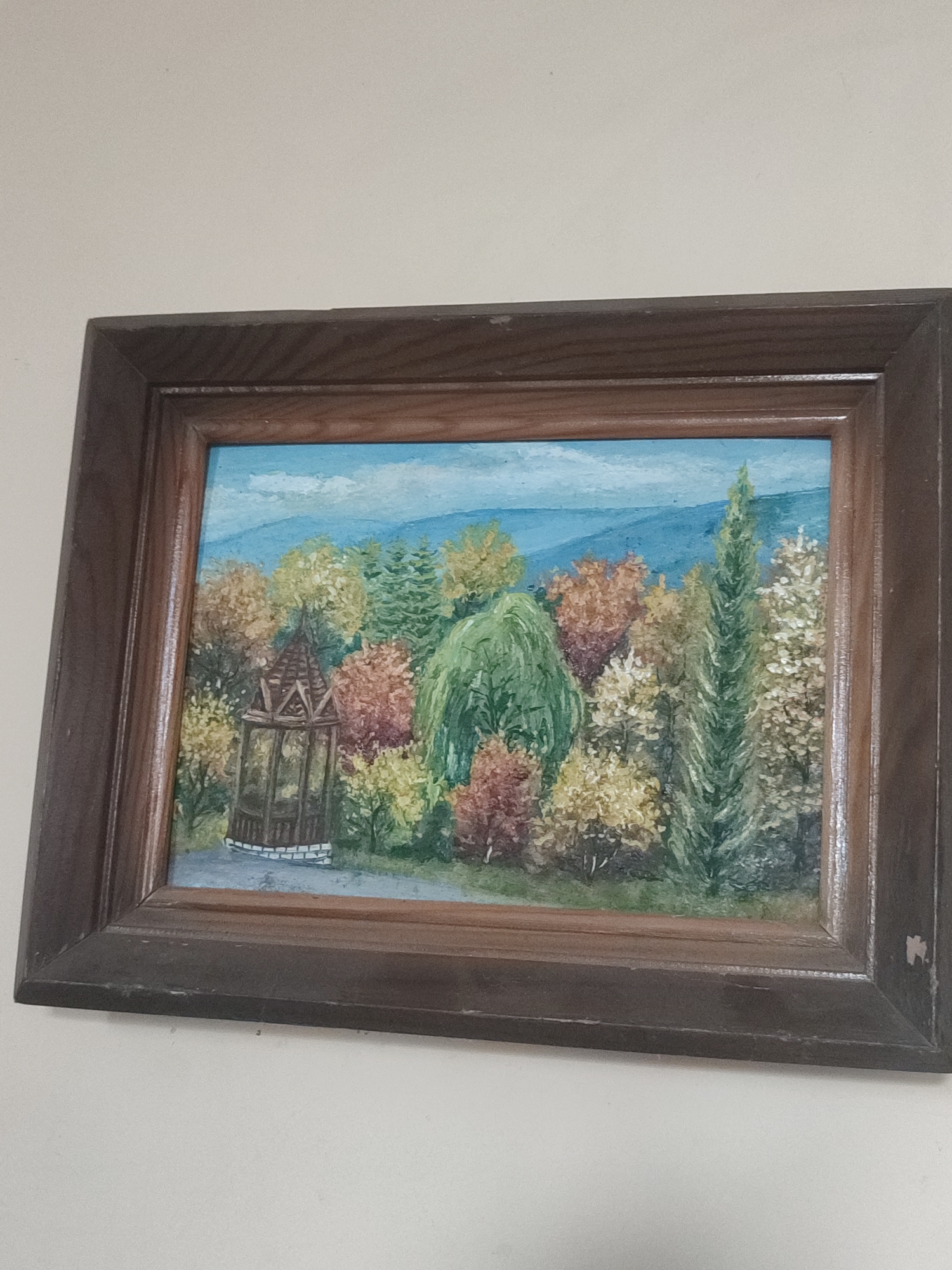 An oil painting in a dark wood frame depicts a scenic landscape. In the foreground, a small gazebo with a brown roof and latticework sides stands on a cobblestone base. Surrounding the gazebo are trees with leaves in various shades of green, yellow, orange, and red, suggesting an autumn or spring setting. A path leads away from the gazebo into the trees. In the background, rolling hills are depicted under a pale blue sky. The painting appears to be a representation of a park or garden. The location of this painting is not specified in Kriukivshchyna, Ukraine, nor is there any information about people or actions occurring within the scene.
